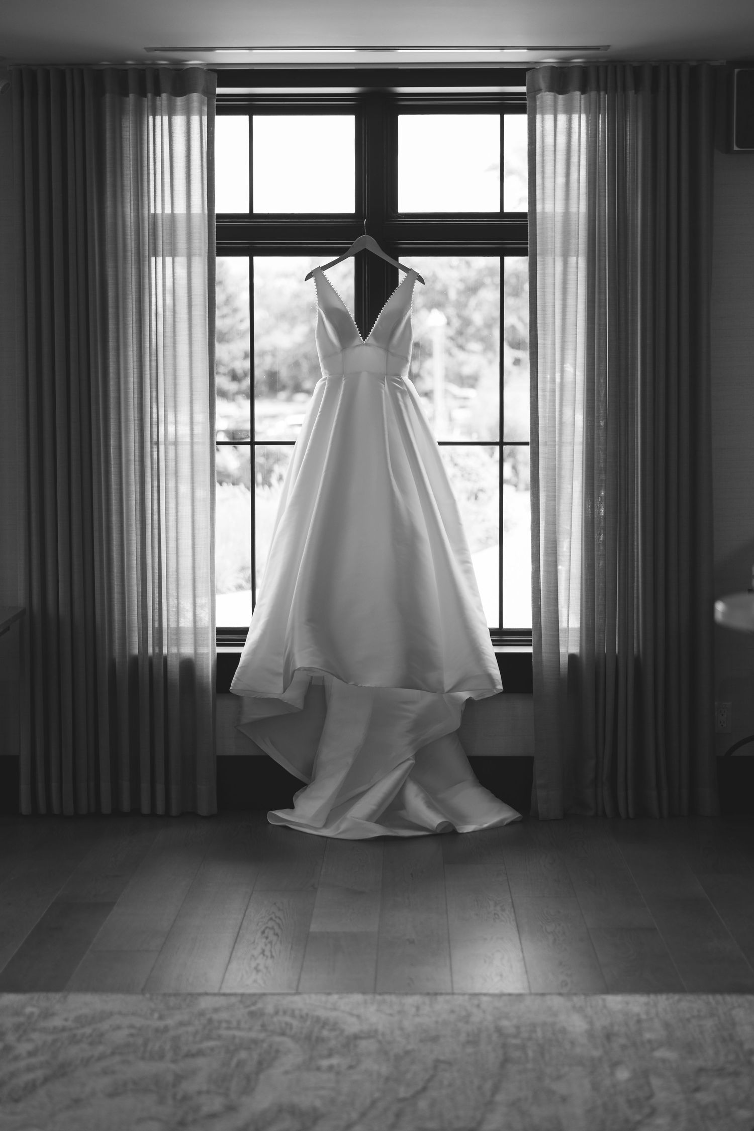 White satin wedding gown with a V-neckline hanging in a sunlit window between sheer curtains, in black and white.