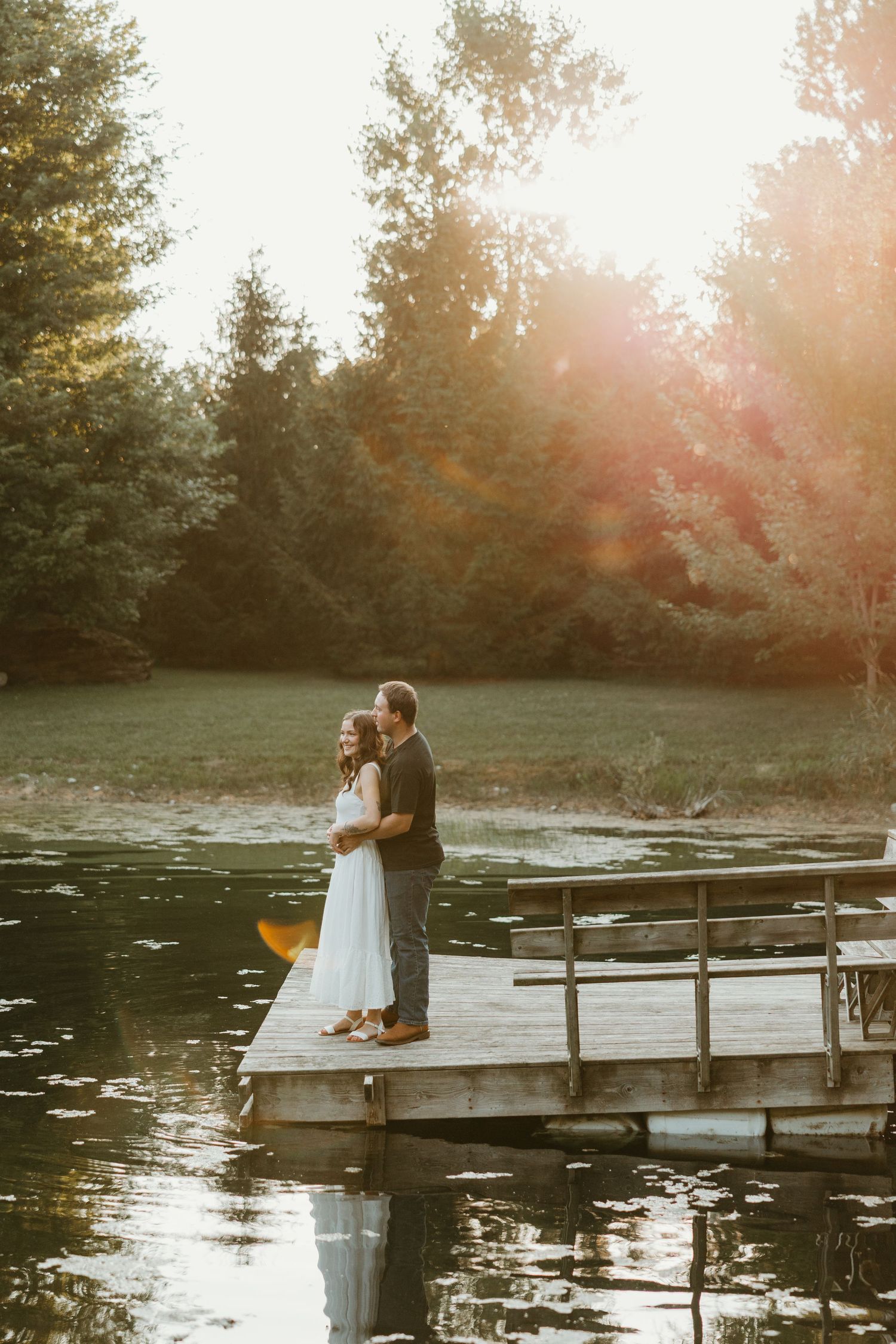 Couple embracing on a wooden dock by a serene lake surrounded by trees at golden hour.