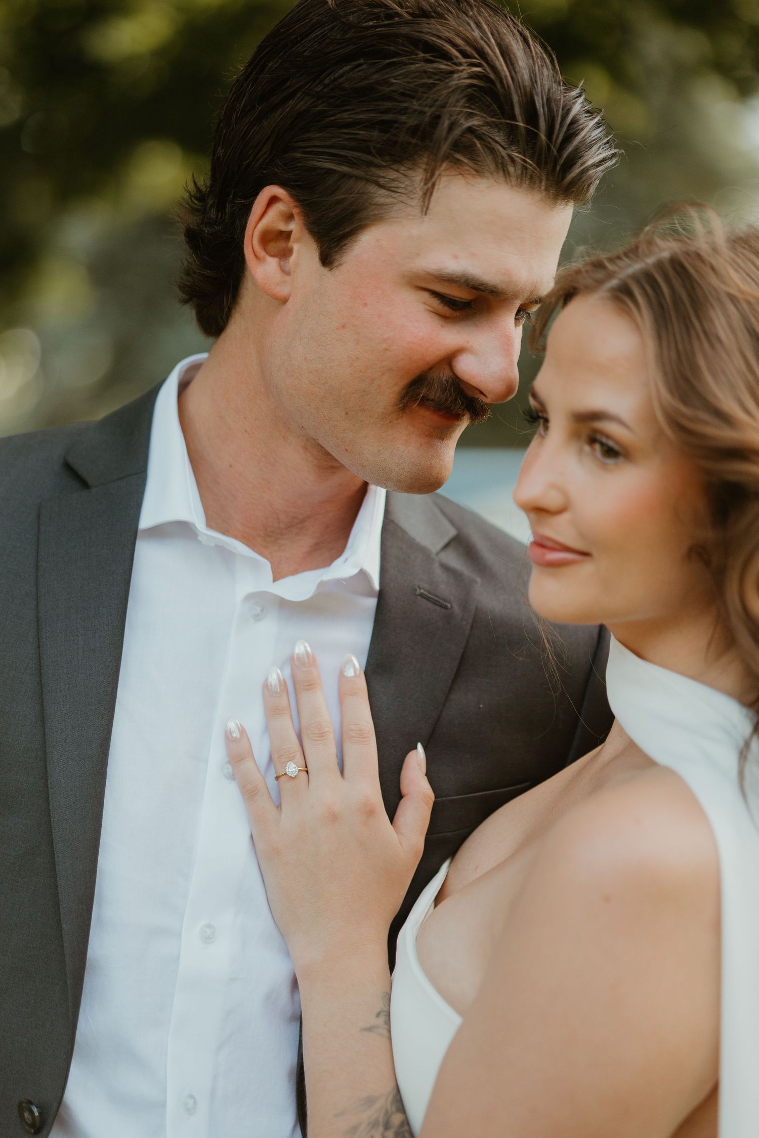 Close-up of smiling bride and groom, bride displaying engagement ring while leaning against groom outdoors.