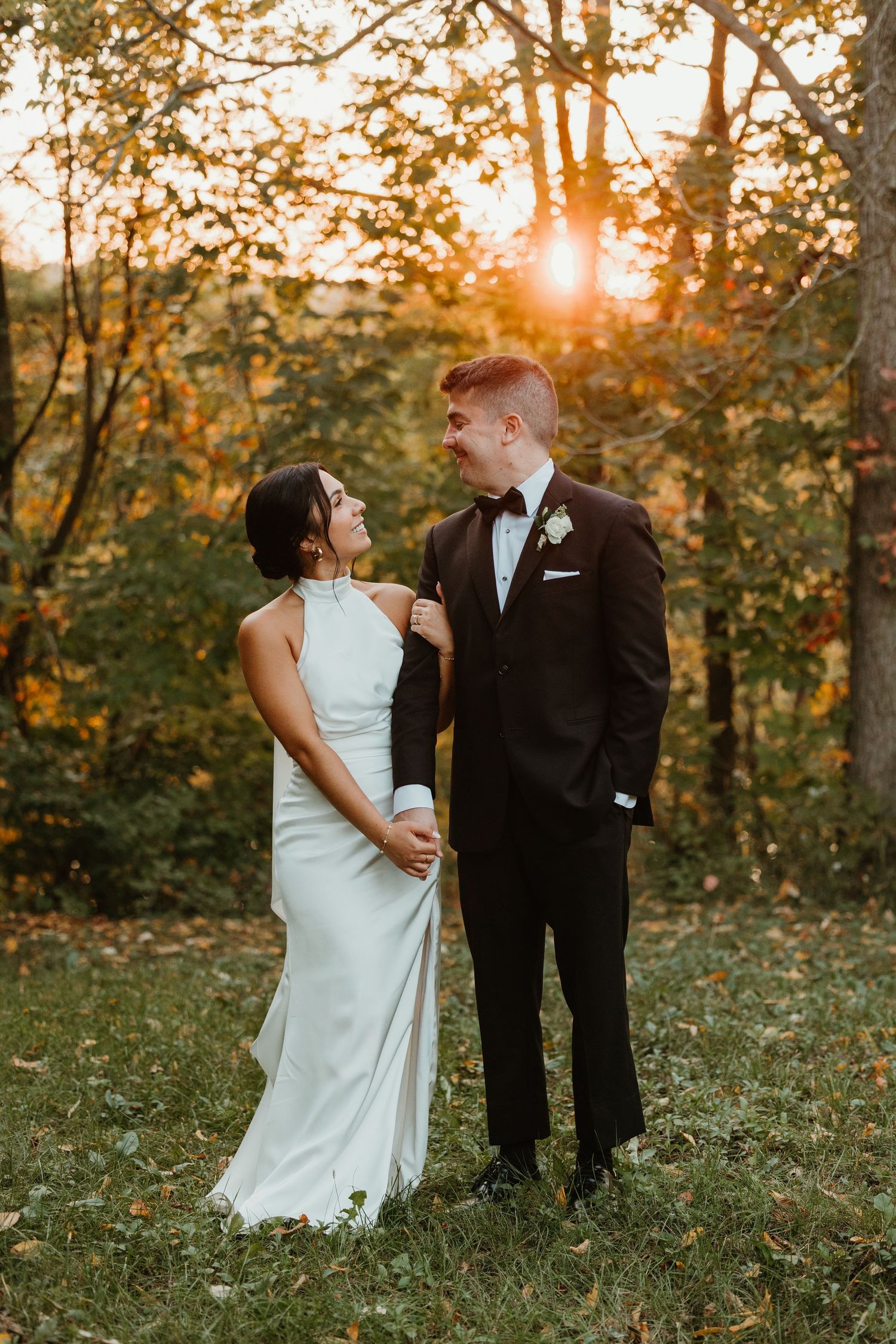 Wedding couple holding hands at golden hour in autumn forest, bride in halter gown, groom in black tuxedo.