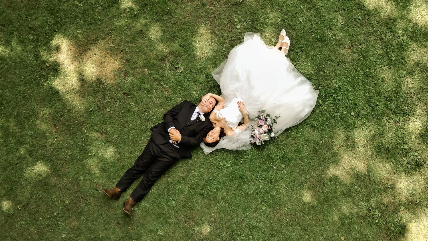 Aerial view of a bride in a white gown and a groom in a dark suit lying together on sunlit green grass.