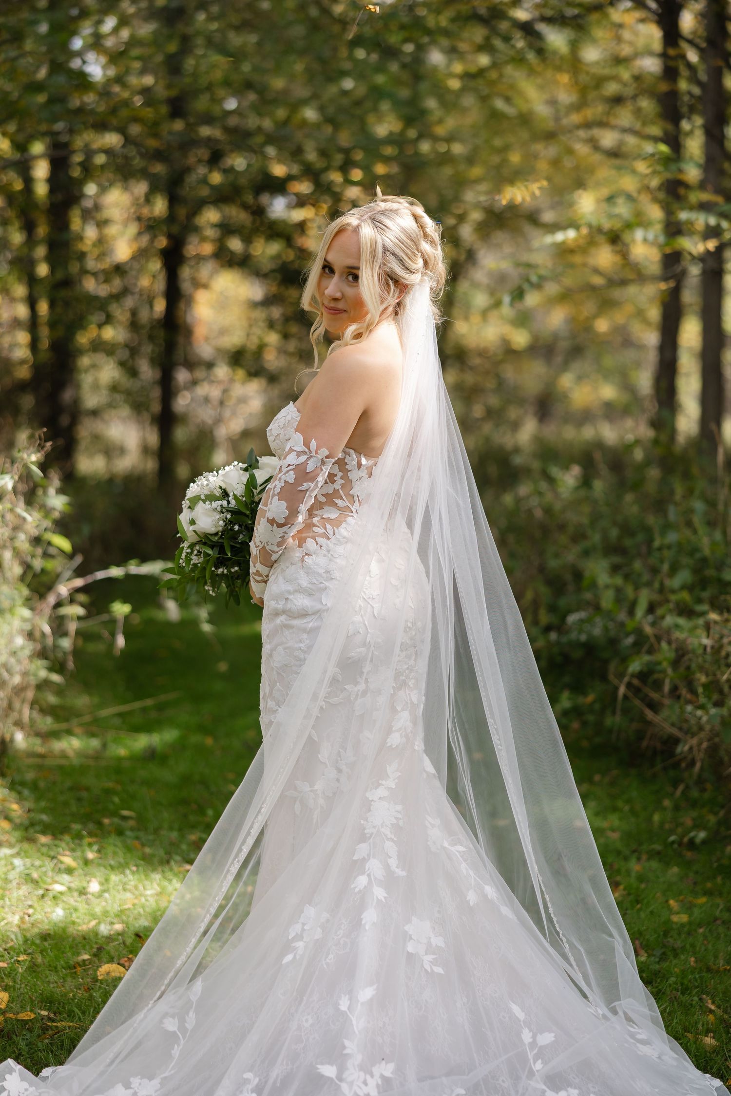 Bride in a lace mermaid wedding gown with a long cathedral veil holding a white bouquet in a sunlit autumn forest.