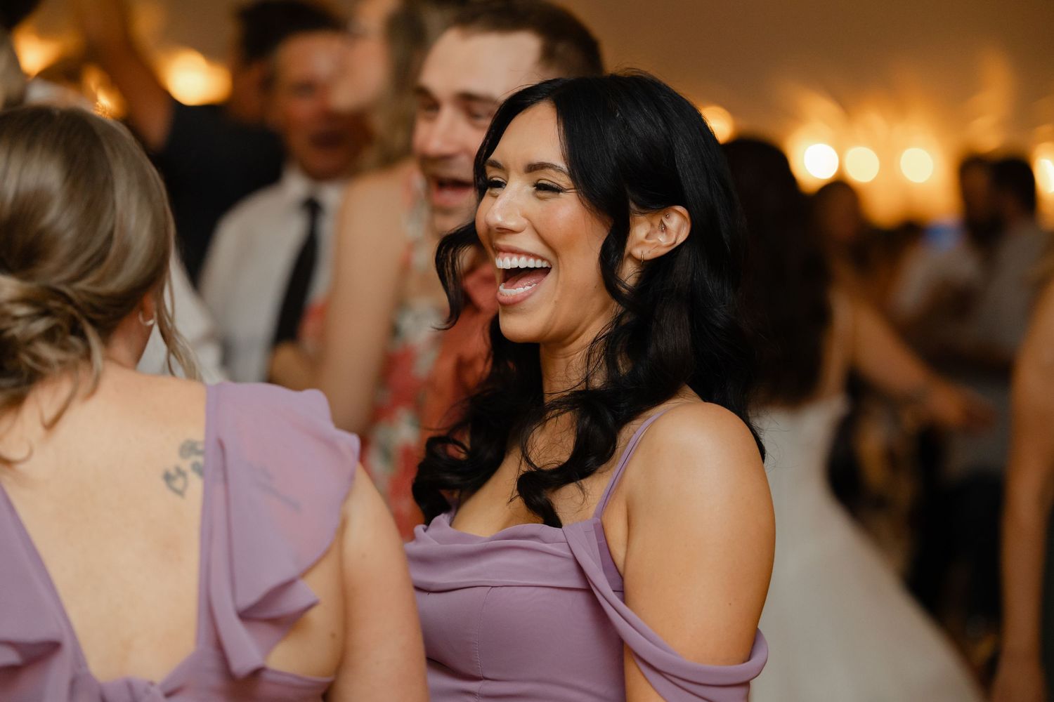 Bridesmaid in lavender off-shoulder dress laughing joyfully with guests on wedding reception dance floor.