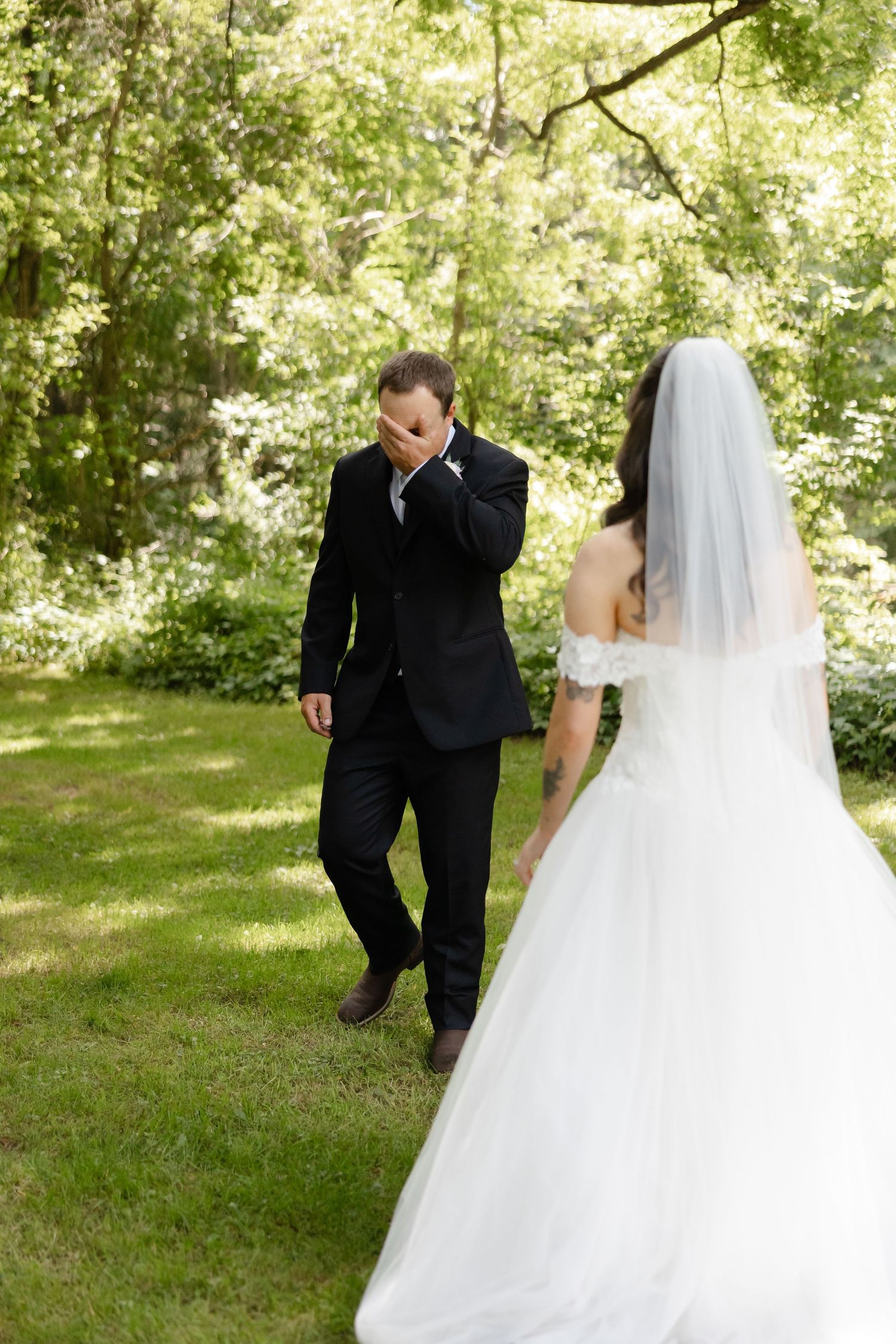 Emotional groom wiping tears during first look with bride in white wedding gown in garden setting.
