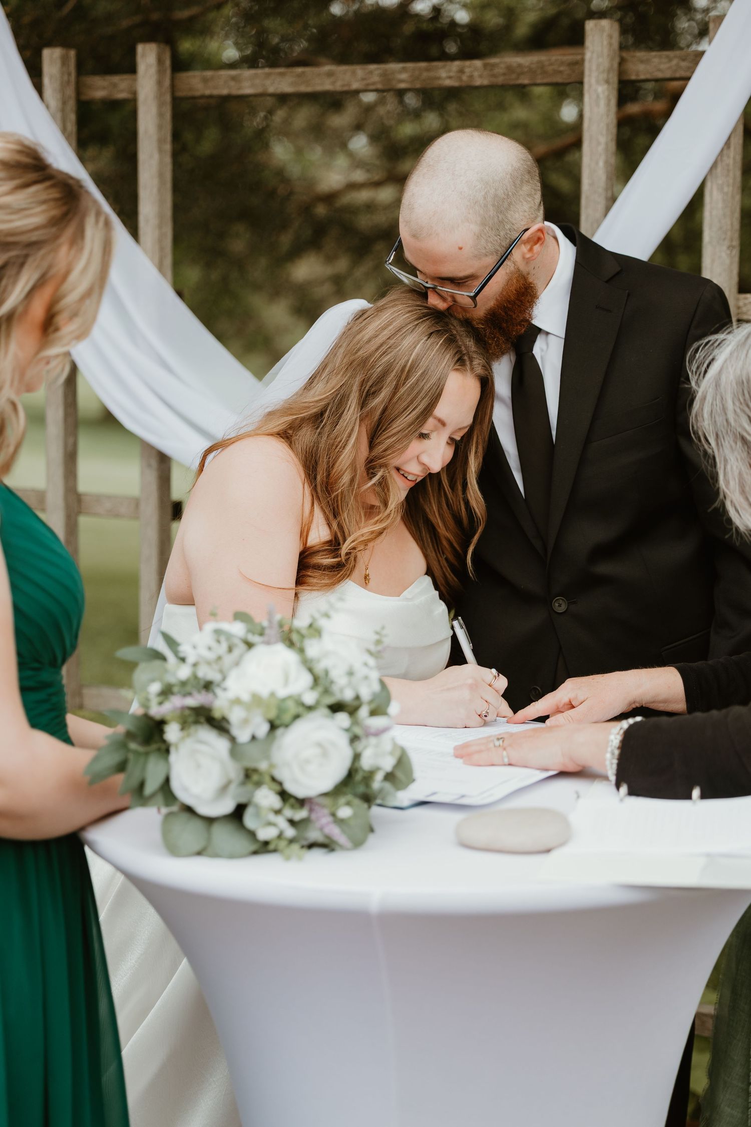 Smiling bride signs wedding documents at outdoor ceremony altar with groom leaning in beside her.