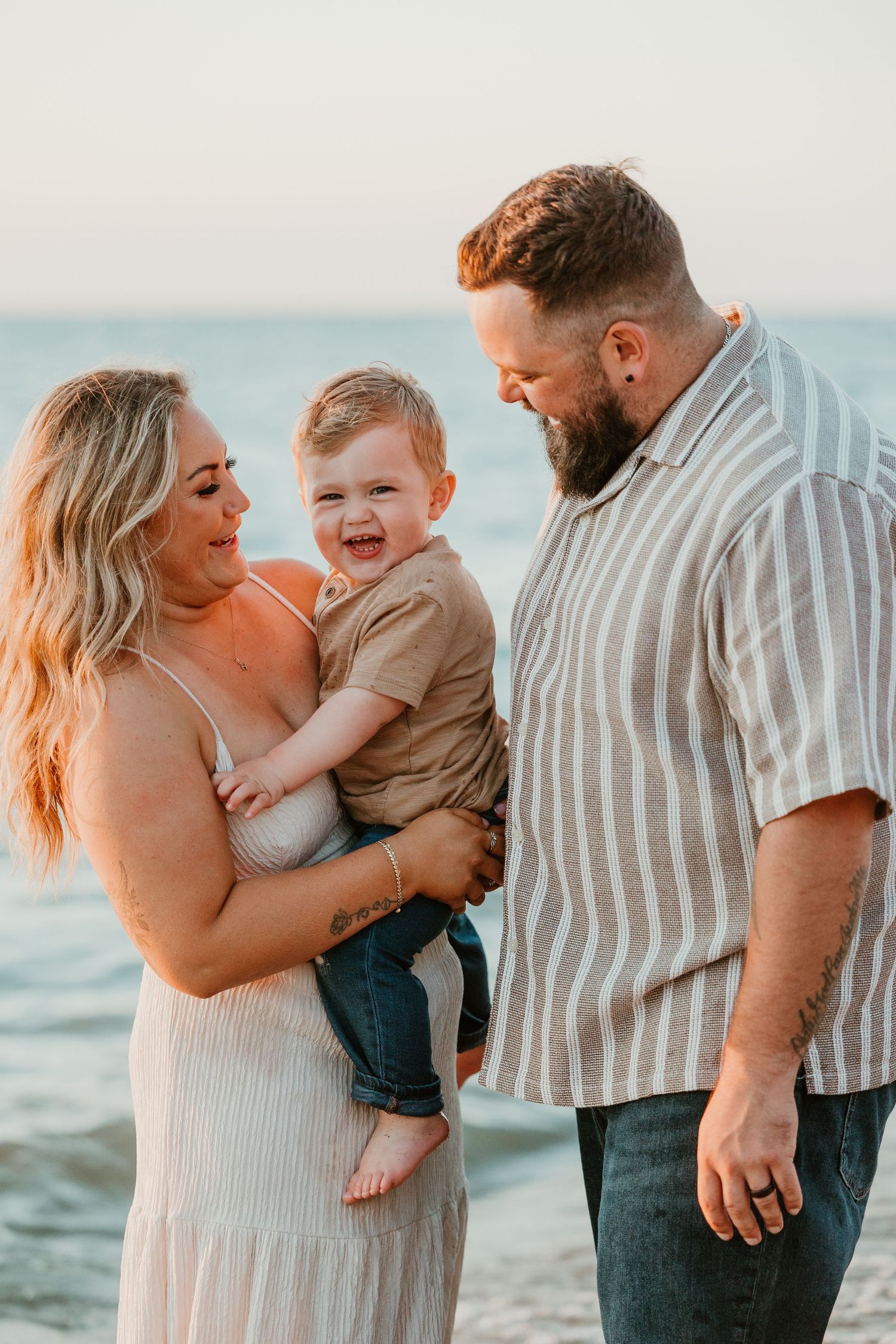 Happy family of three at golden hour beach, parents smiling at giggling baby boy held between them.