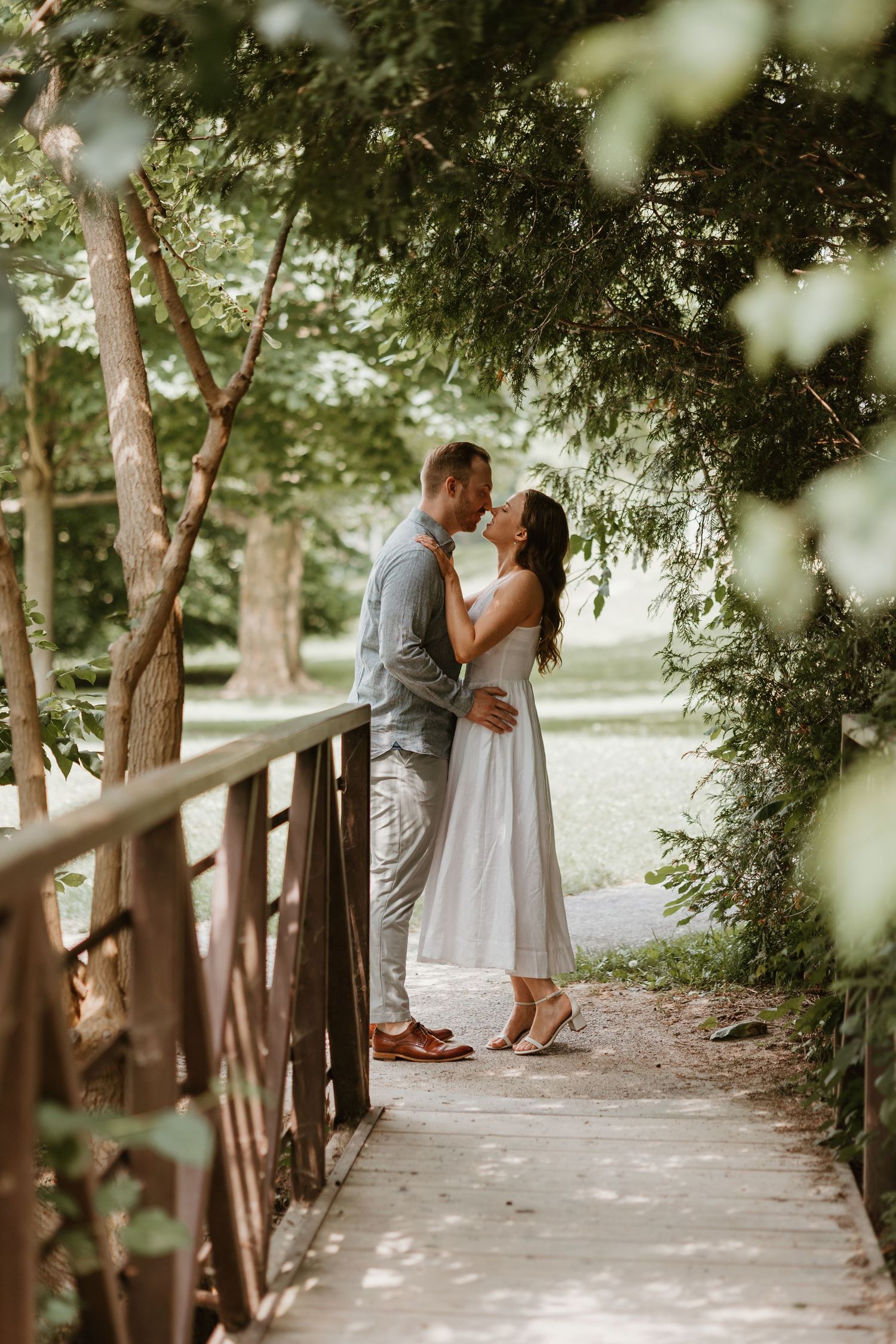 Couple sharing a romantic kiss on a wooden bridge surrounded by lush green trees.
