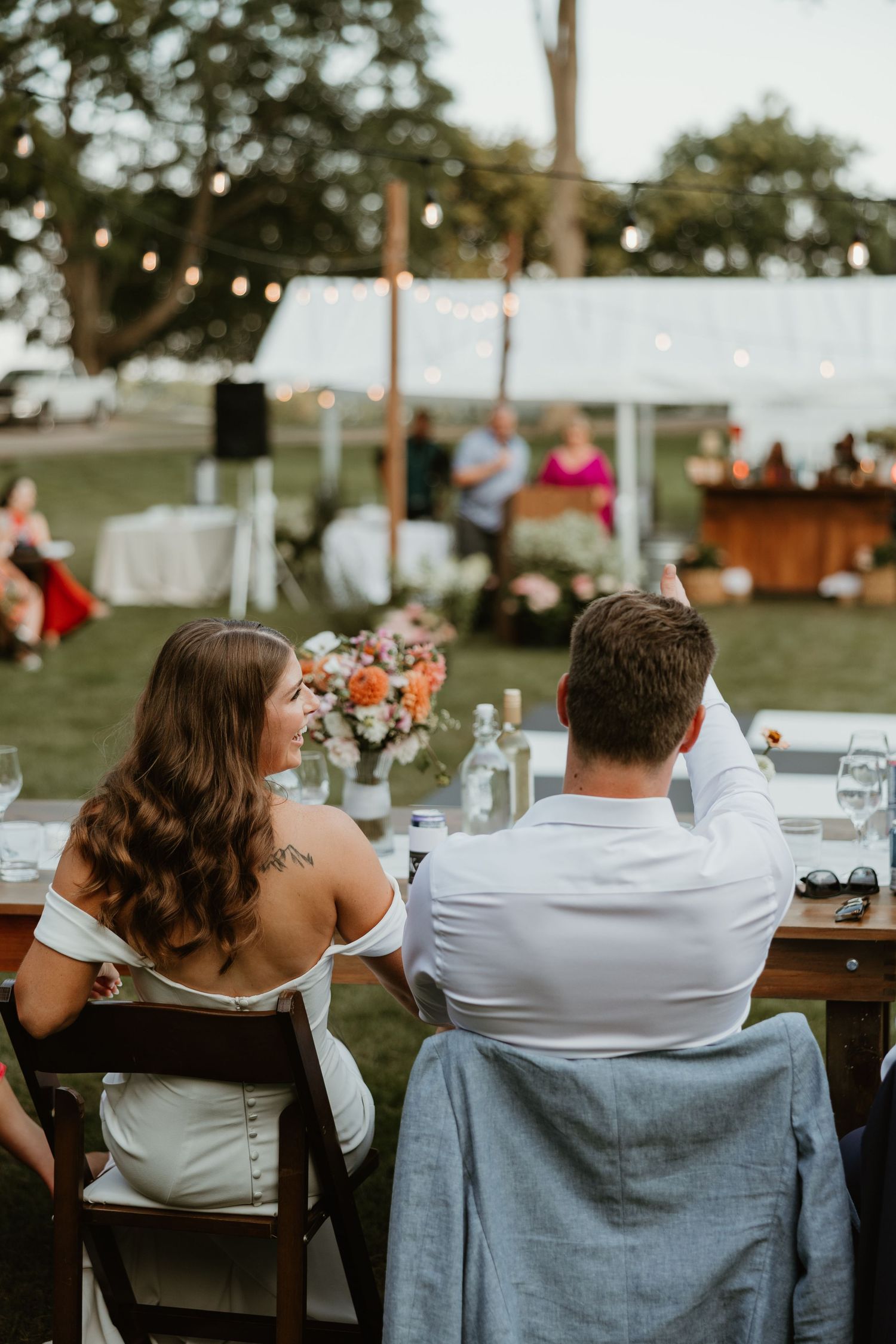 Bride and groom seated at outdoor reception table with string lights and colorful floral centerpiece at dusk.