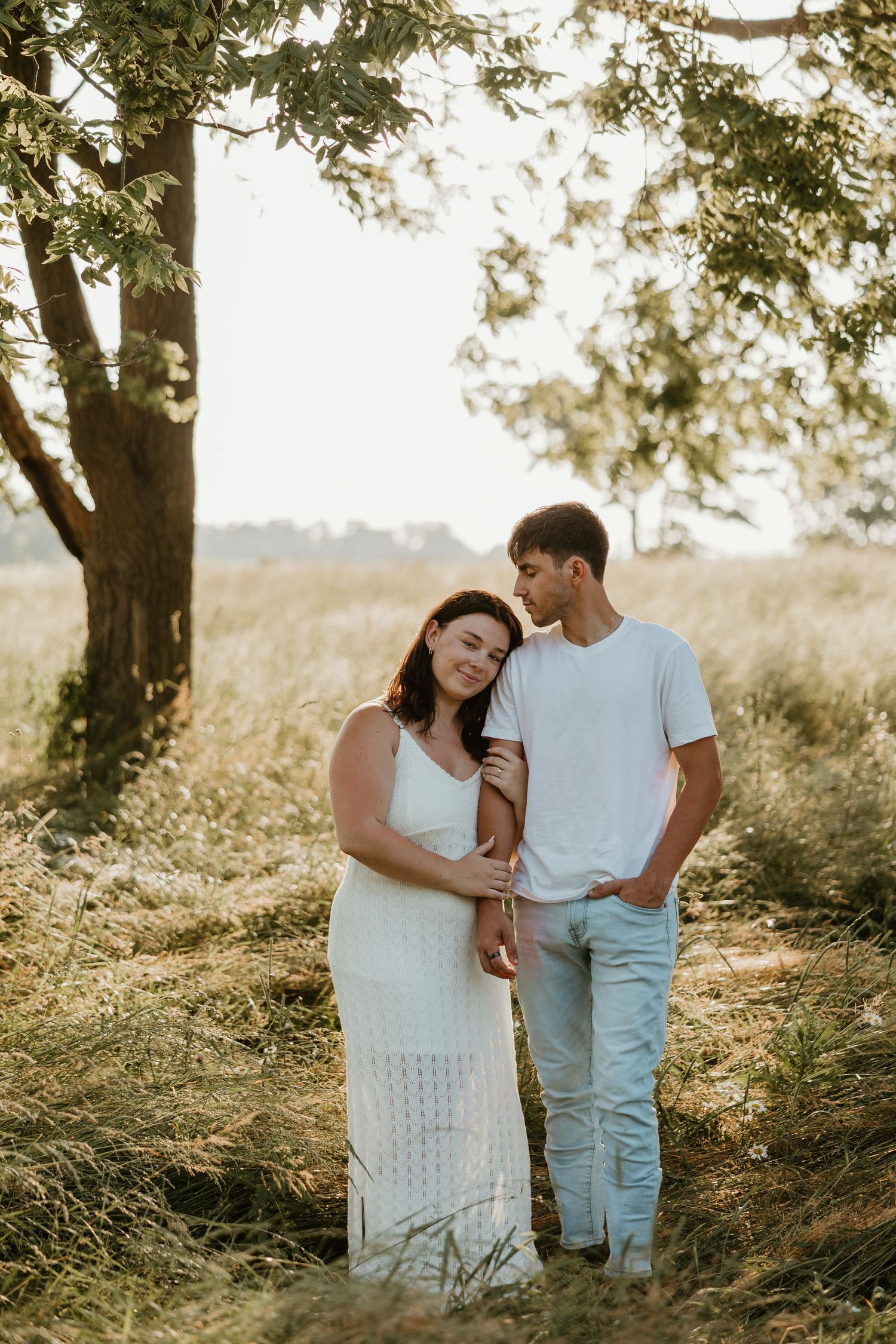Couple embracing under a tree in a sunlit field, woman in white dress leaning on mans shoulder.