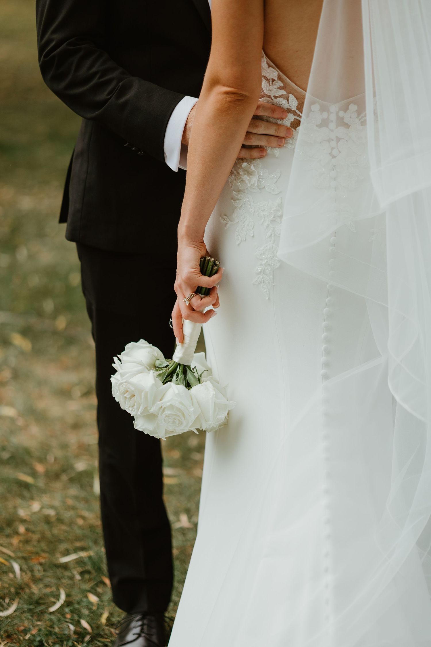 Close-up of bride in lace wedding gown holding white rose bouquet with groom in black suit.