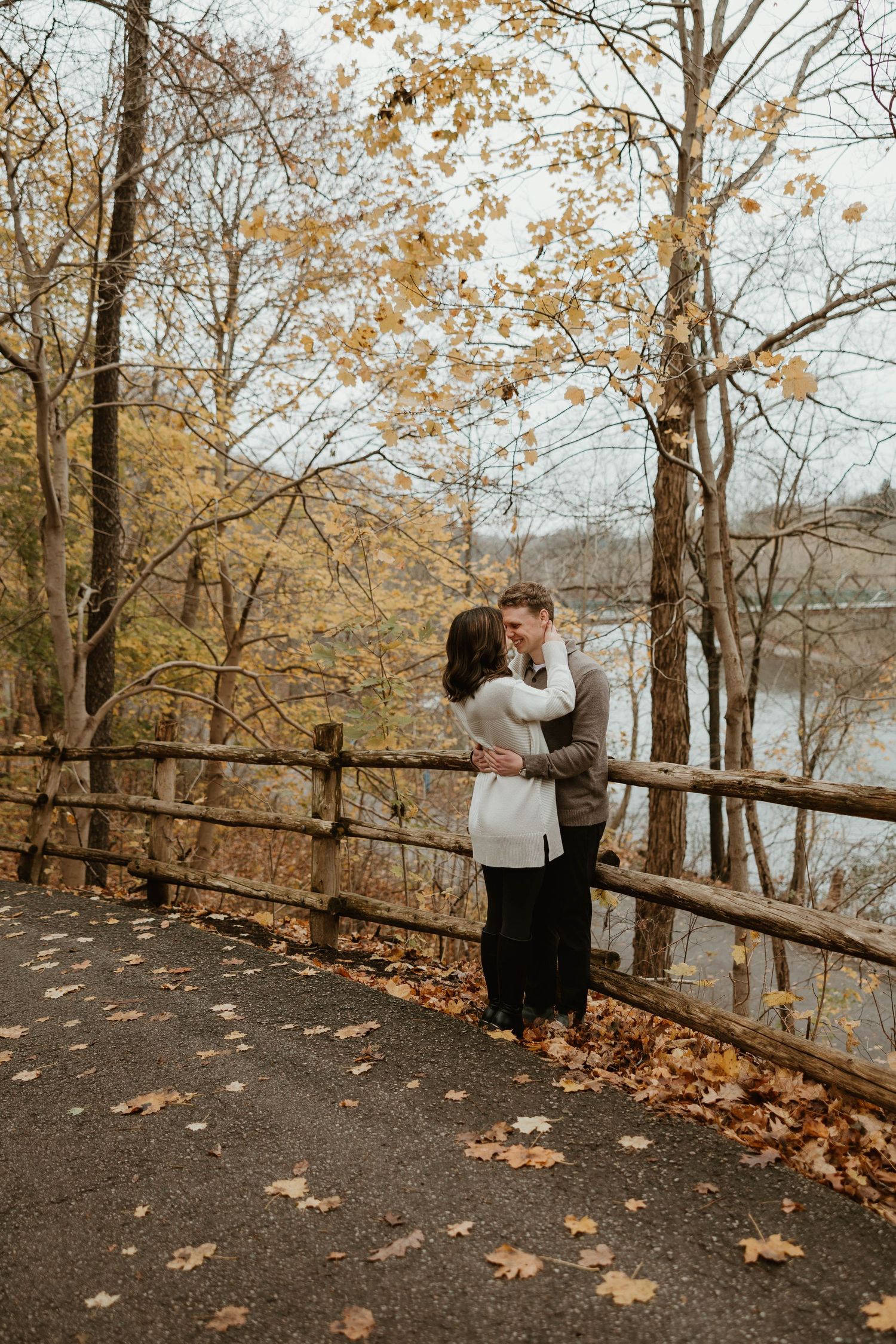 Romantic couple embracing near wooden fence by lake in fall foliage setting.