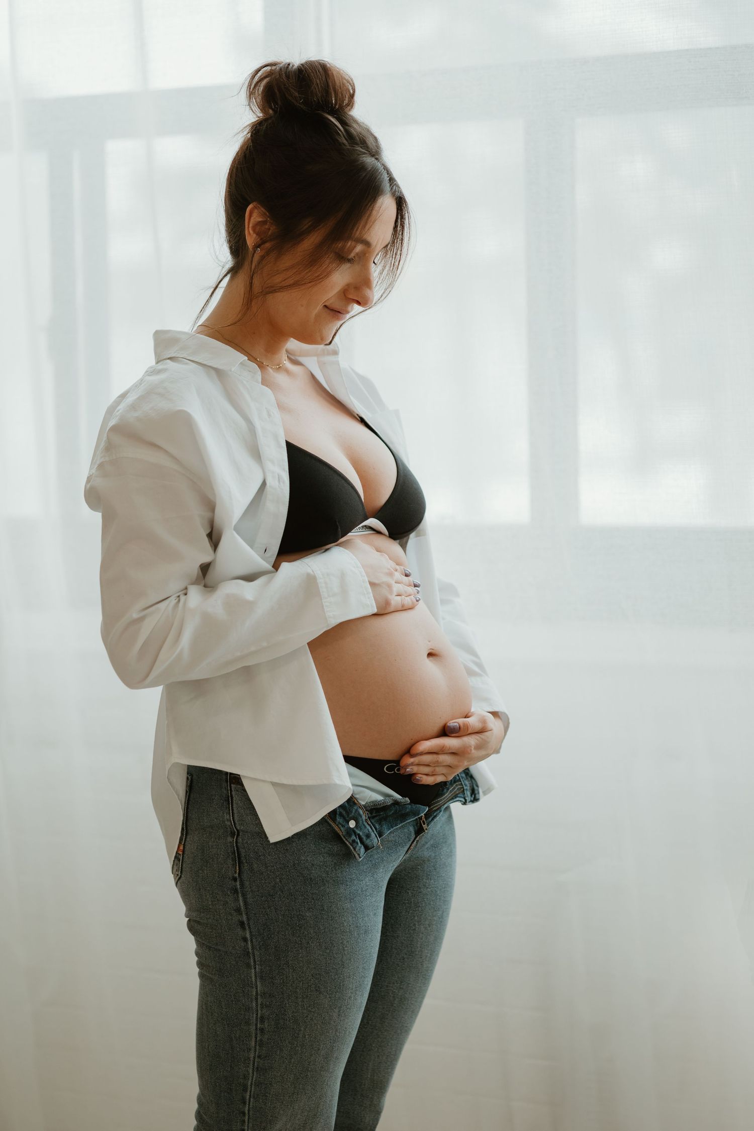 Pregnant woman in black bra and white shirt holding her bare baby bump while looking down.