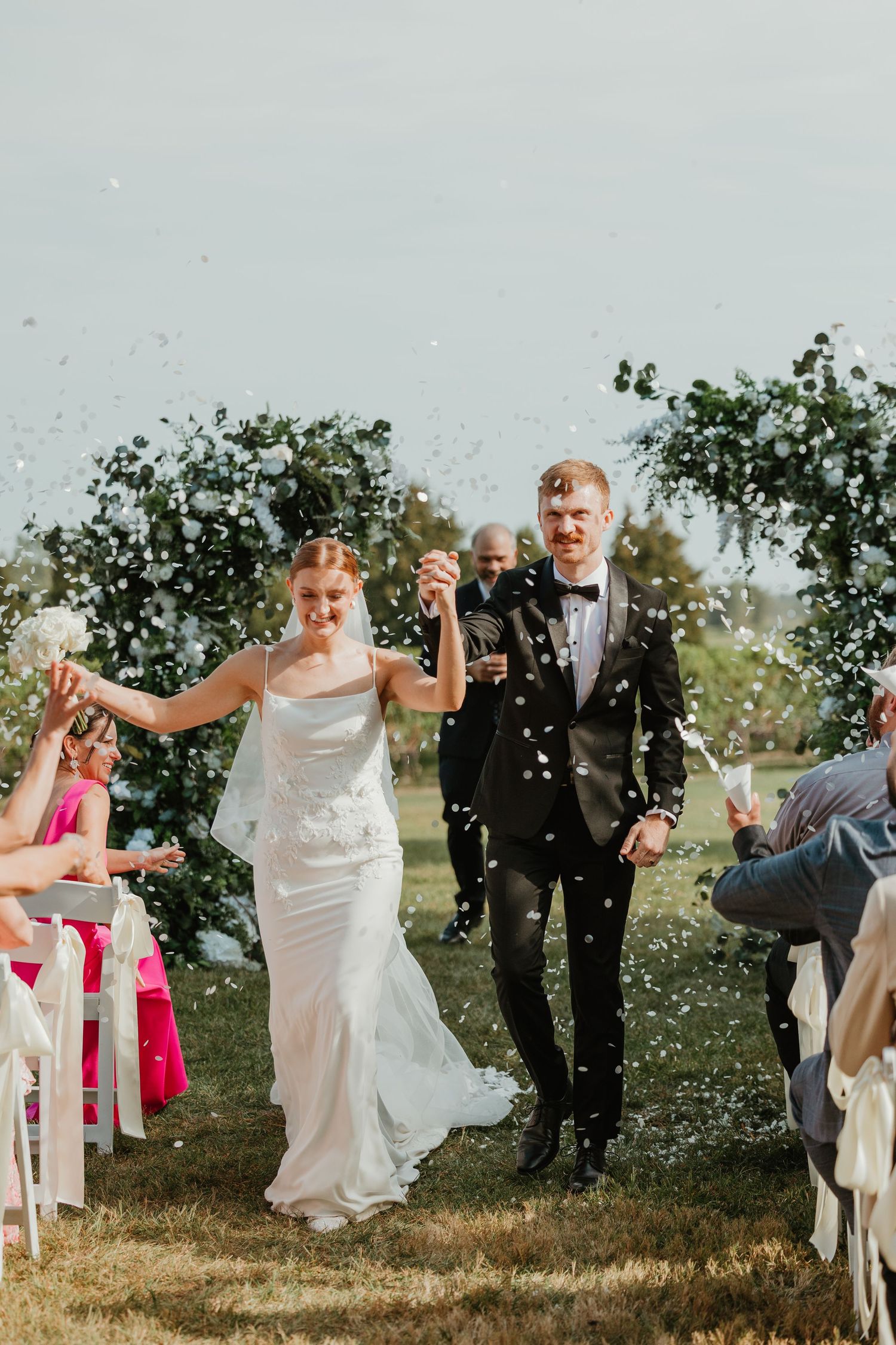 Bride and groom walk down the aisle as guests toss white confetti at outdoor wedding ceremony.
