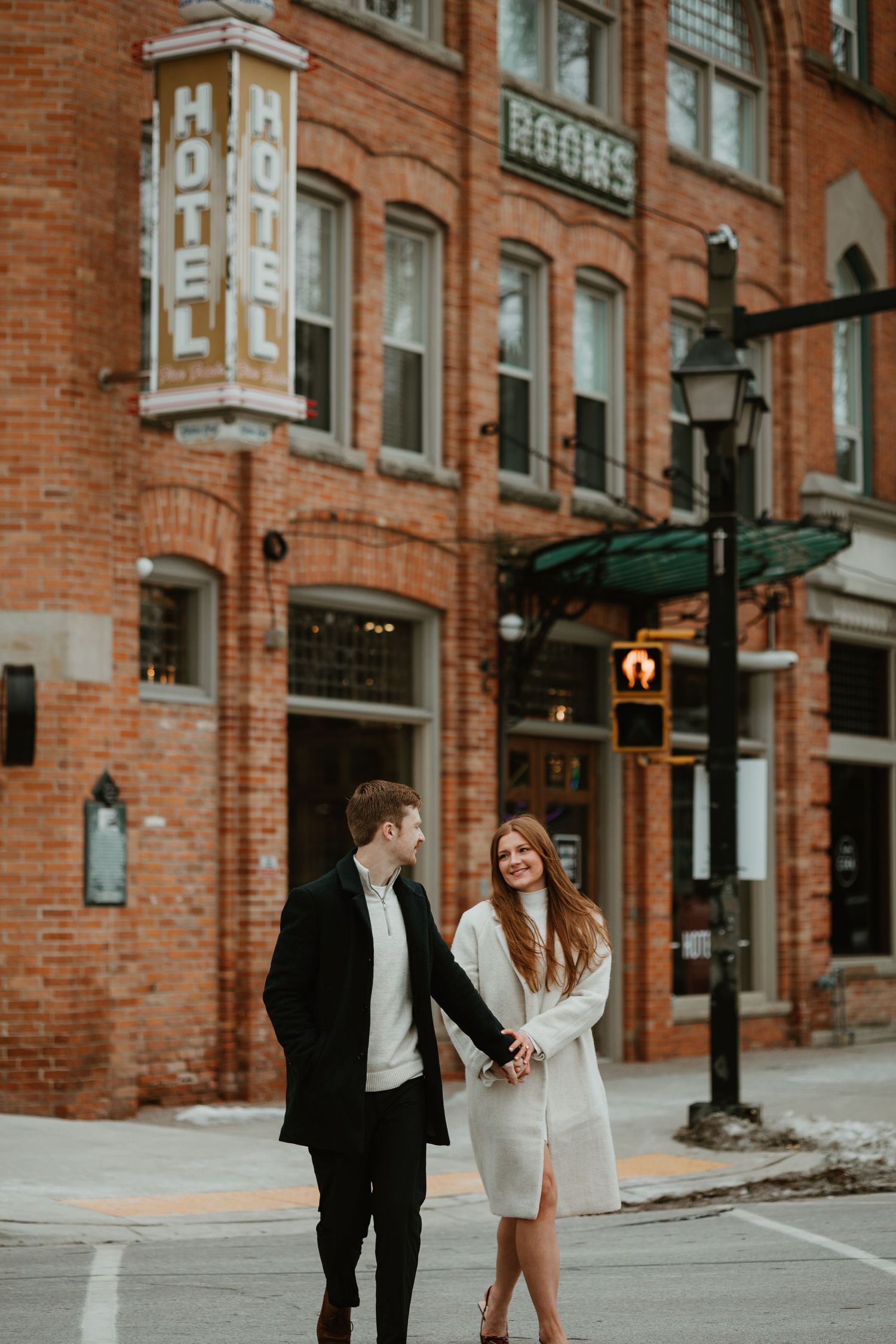 Couple holding hands and laughing while crossing a street in front of a historic brick hotel building.