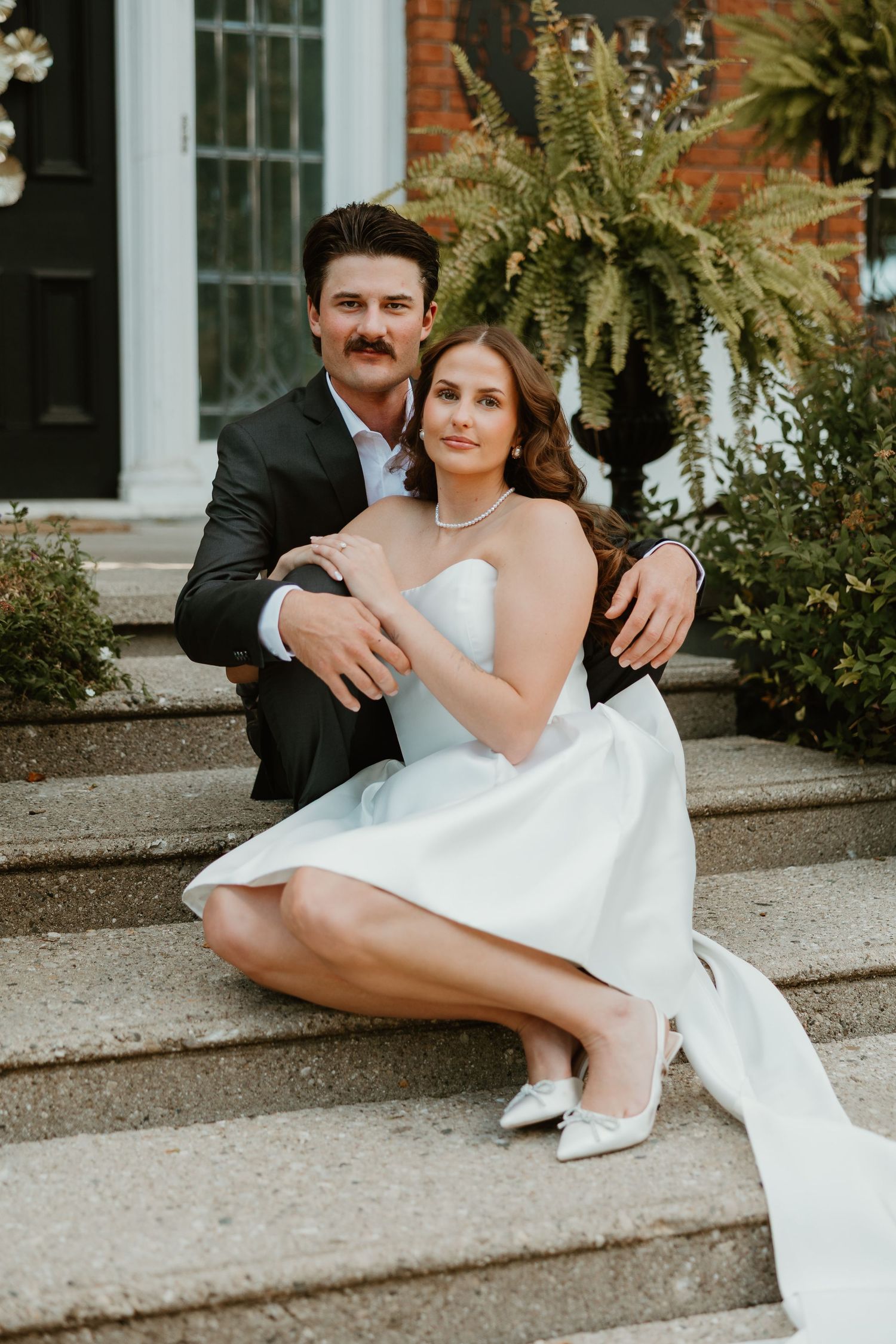 Bride and groom sitting together on stone steps of an elegant home, bride in white strapless dress.