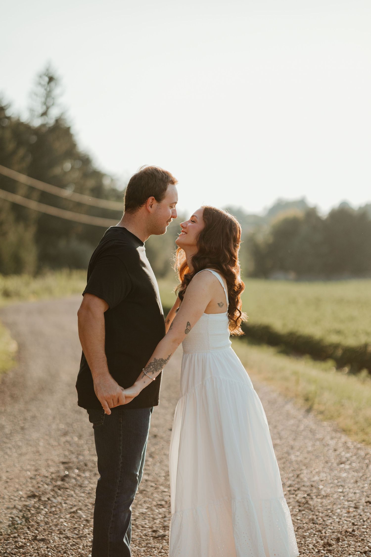 Couple holding hands and smiling at each other on a rural gravel road during sunset engagement shoot.