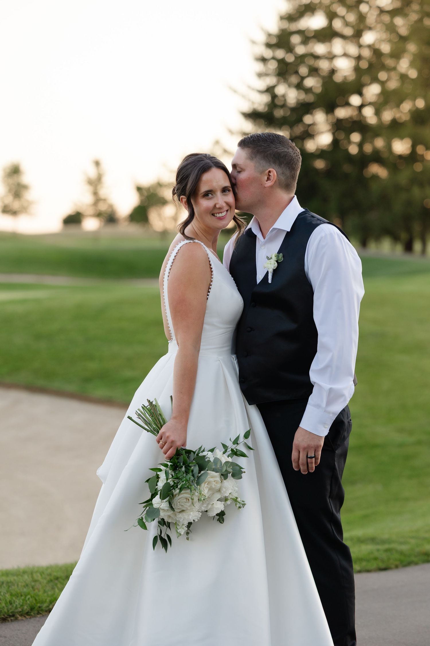 Smiling bride in a white wedding dress holding a white floral bouquet as the groom in a vest kisses her cheek on a golf course.