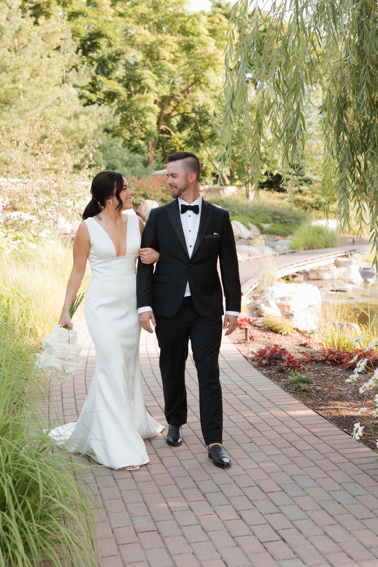Bride in white gown and groom in black tuxedo walk hand-in-hand along a garden brick pathway.