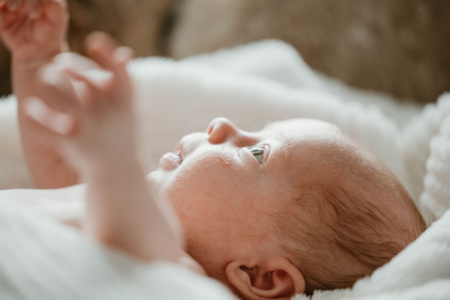 A peaceful newborn baby lies on a white blanket with arm raised in a serene close-up portrait.