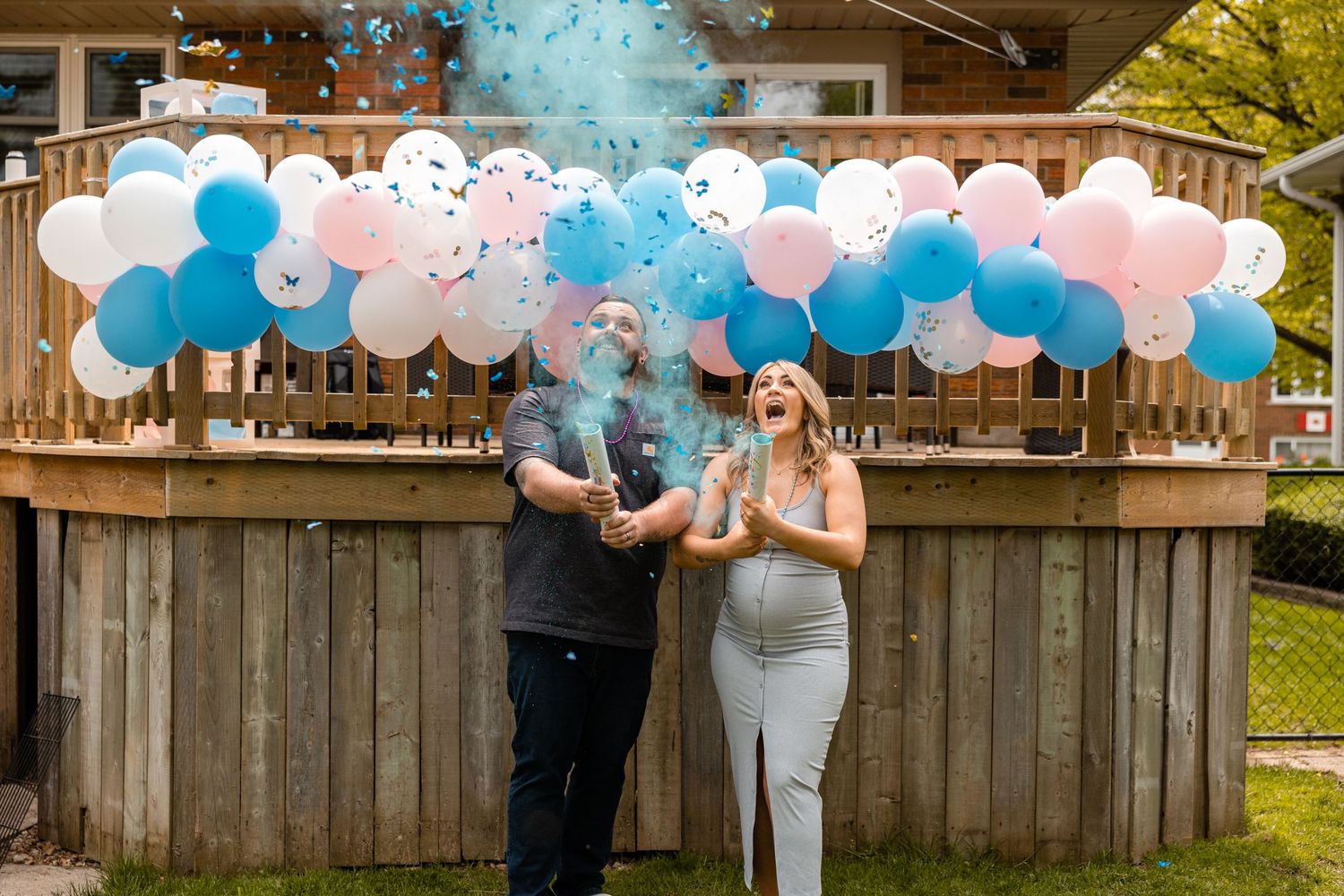 Excited couple at gender reveal with blue confetti bursting into the air by balloon arch.