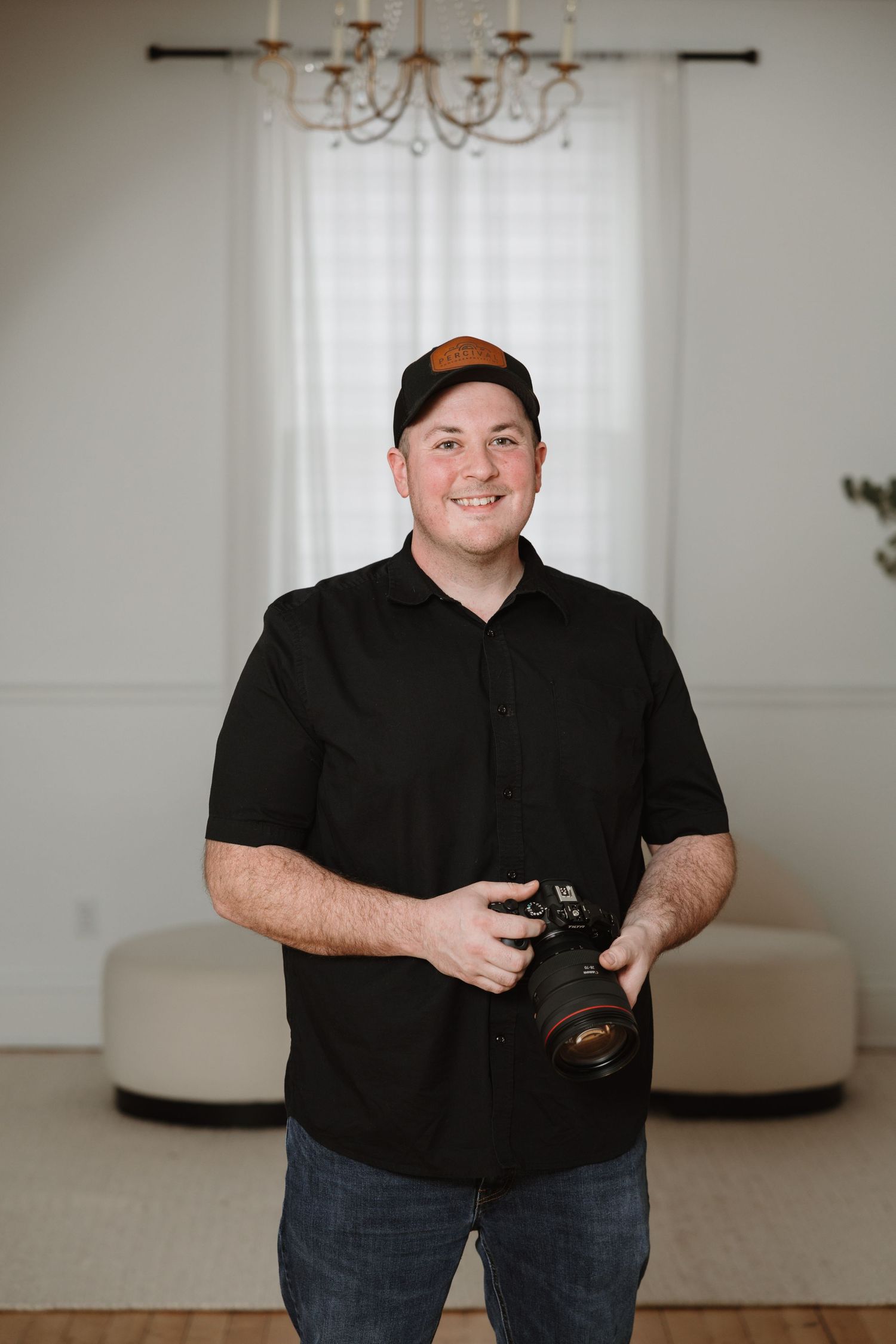 Male photographer smiling and holding camera in bright studio wearing black shirt and cap.
