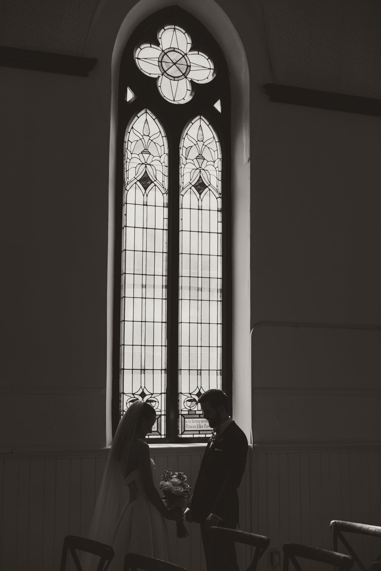 Silhouette of bride and groom holding hands before tall stained glass church window.