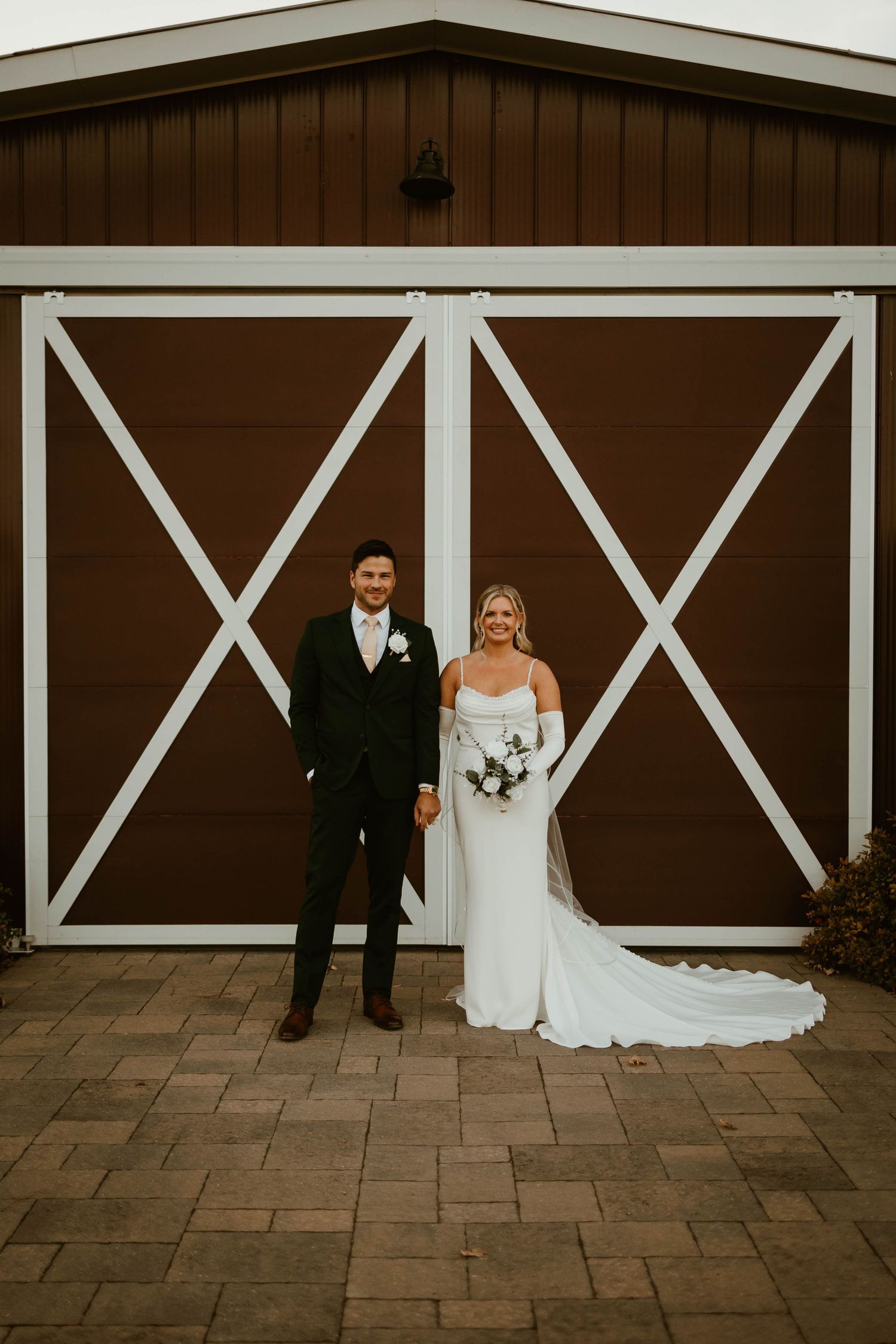 Bride in white gown and groom in dark suit stand before a brown and white barn door.