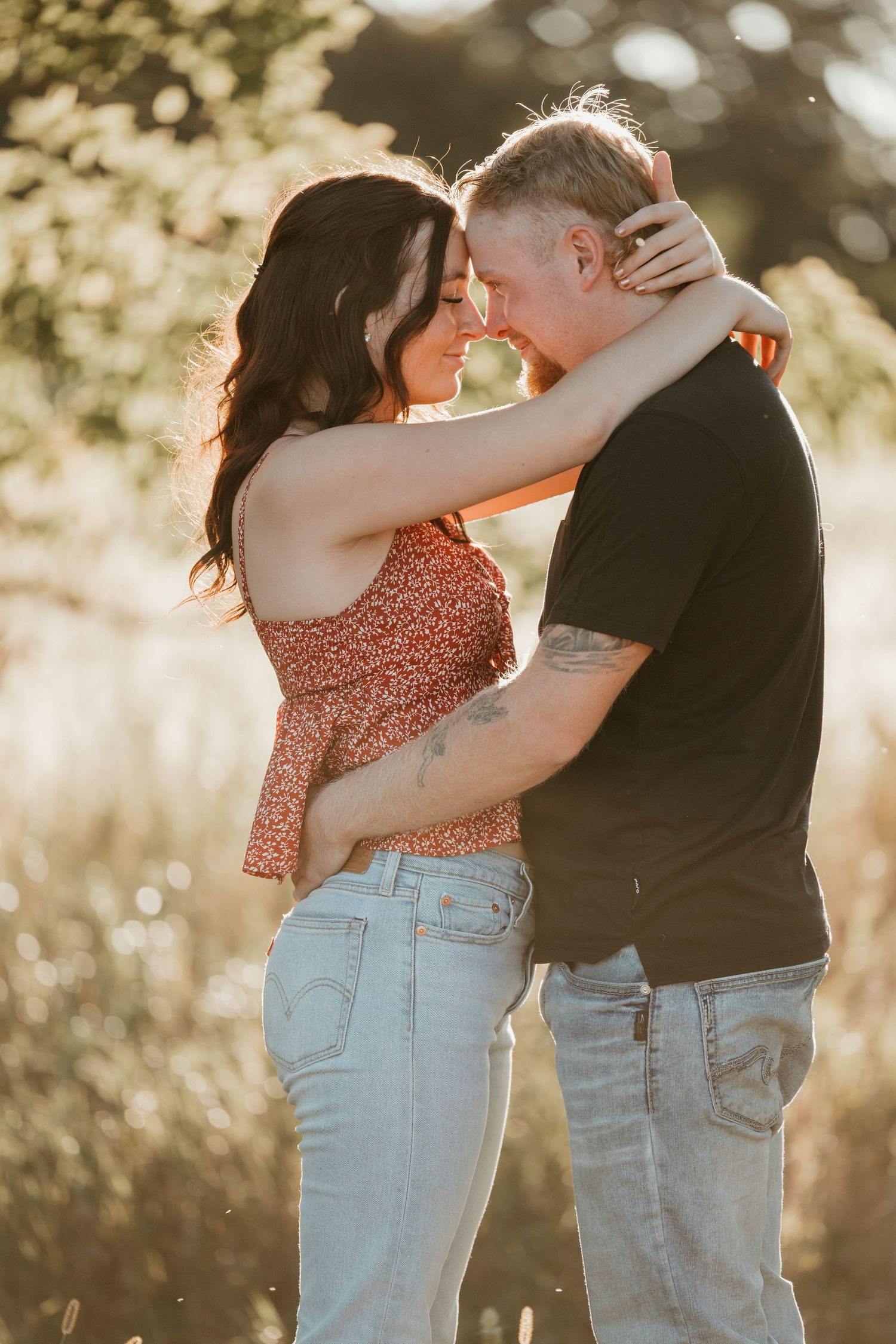 Couple embracing in golden sunlight in field, woman in floral top and jeans.