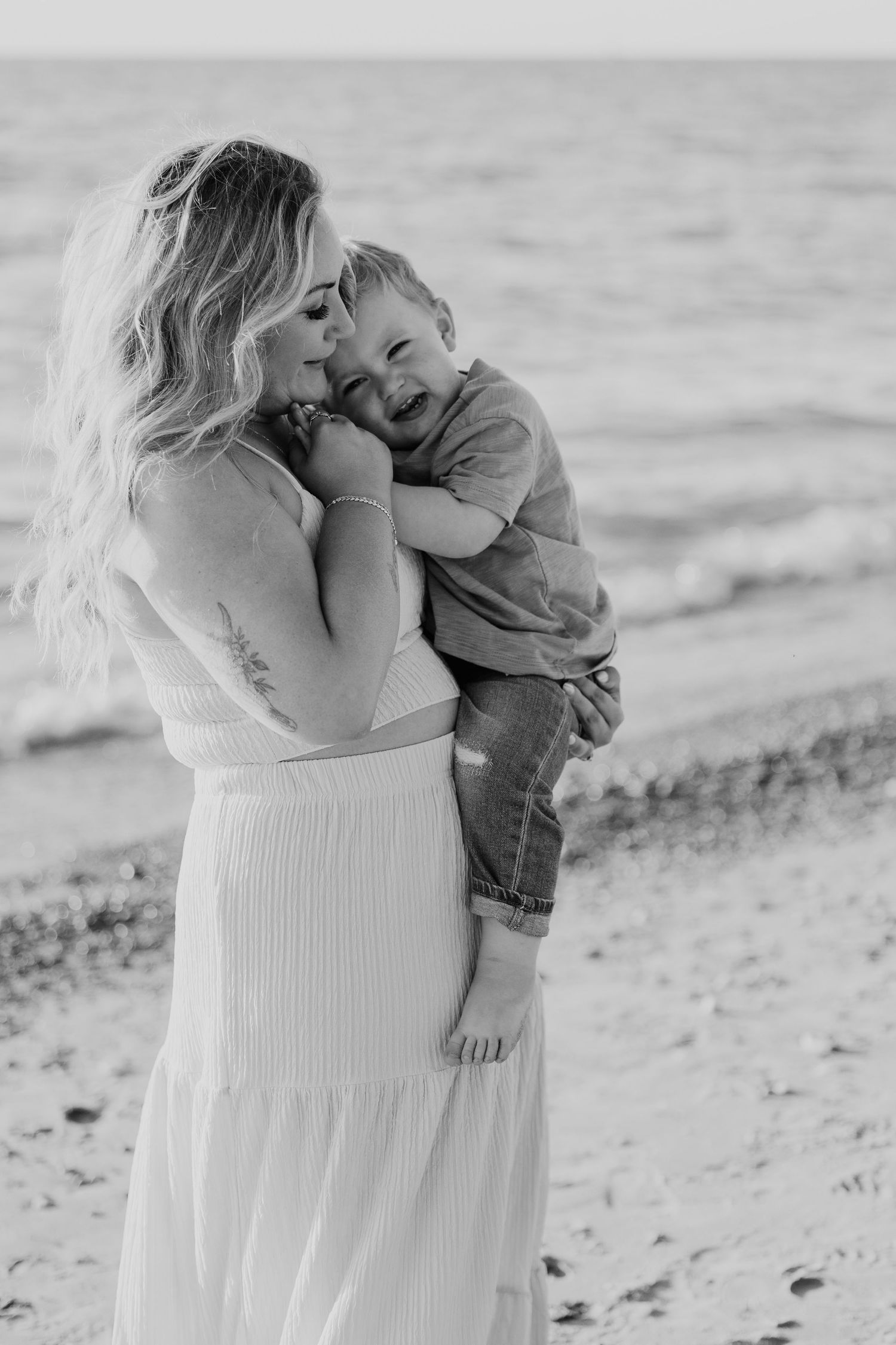 Black and white photo of smiling mother holding laughing toddler on sandy beach with ocean waves behind them.