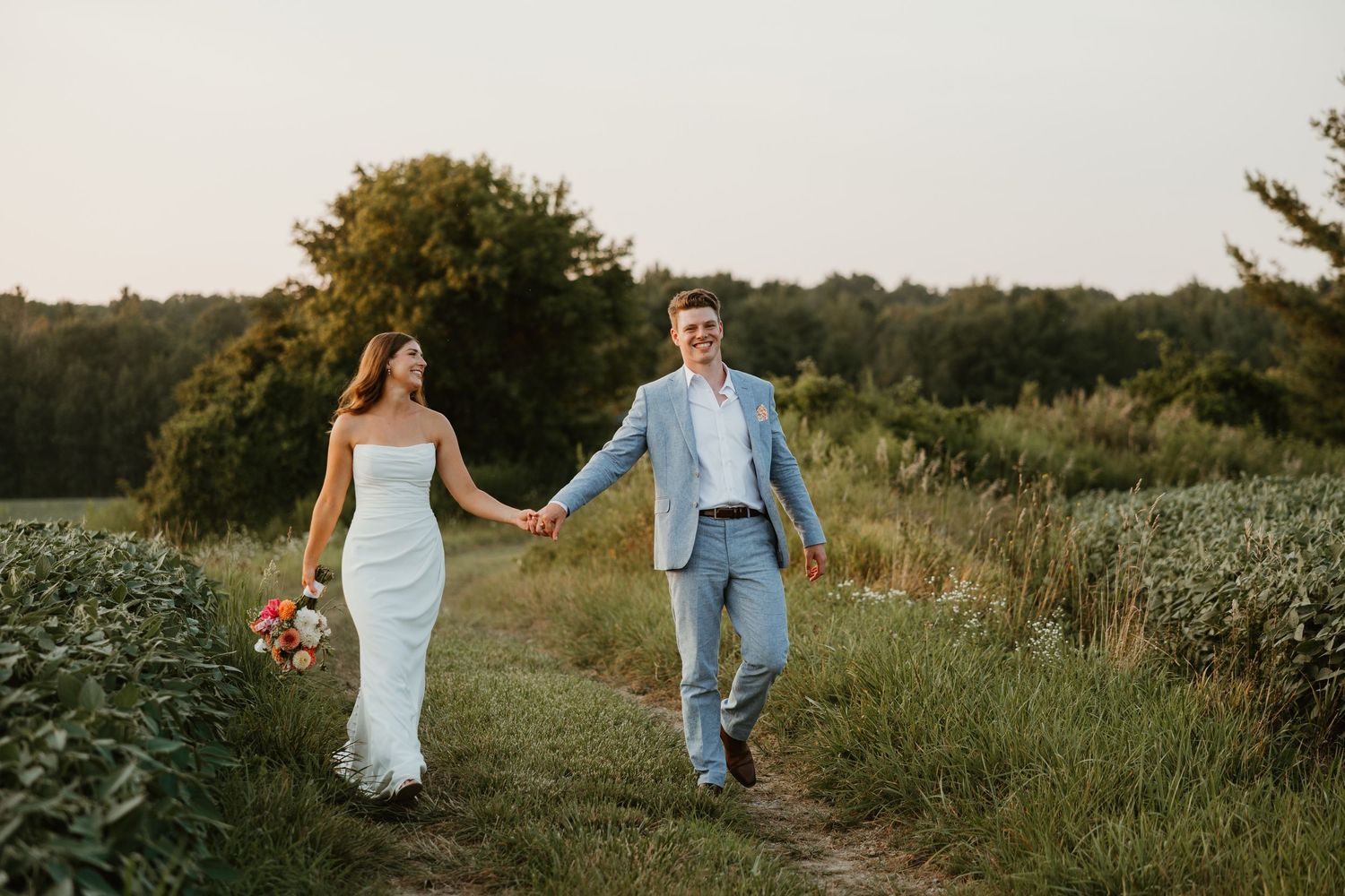 Happy bride and groom holding hands walking along a scenic countryside path at sunset.