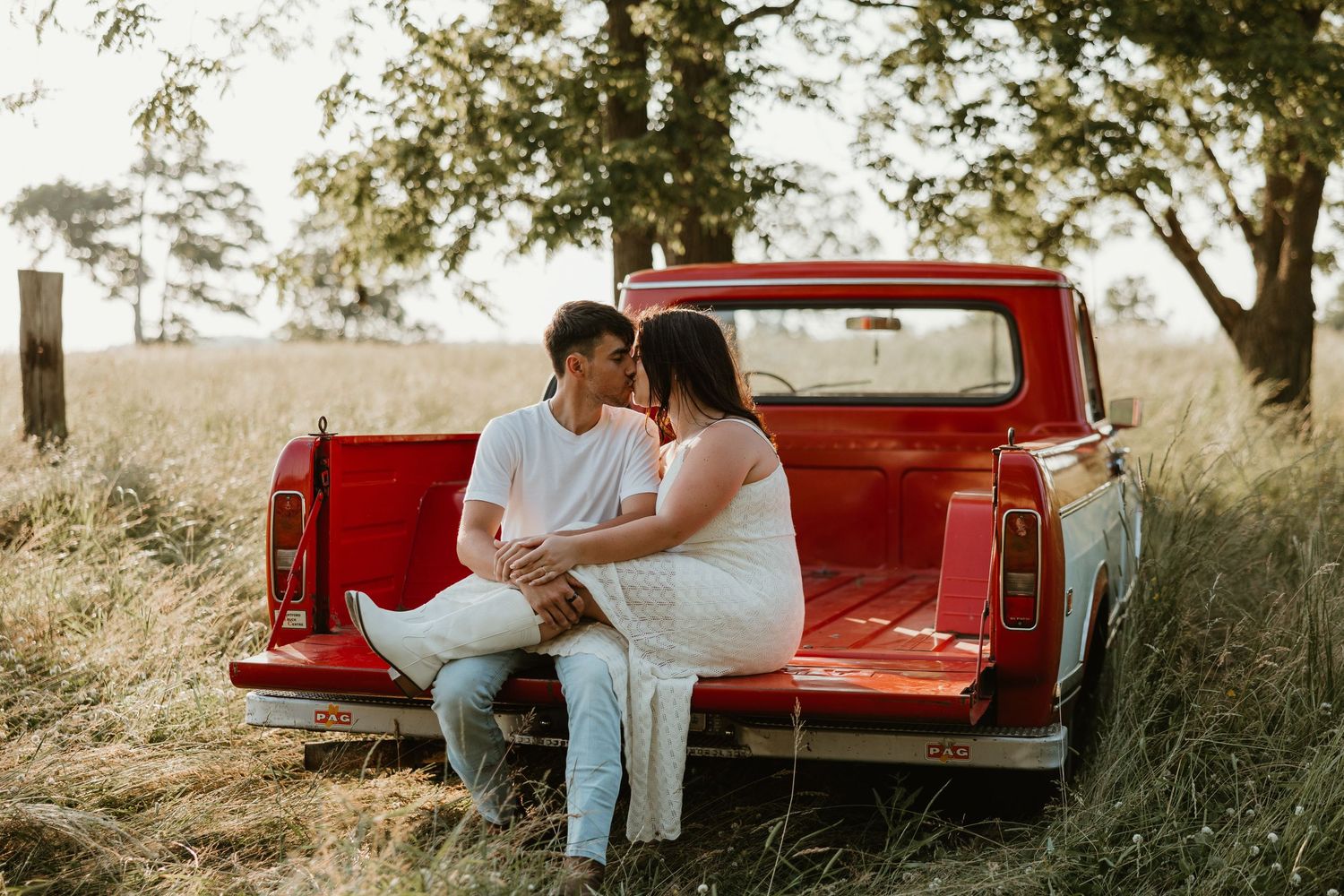Couple sitting closely together on the tailgate of a red vintage pickup truck in a golden field.
