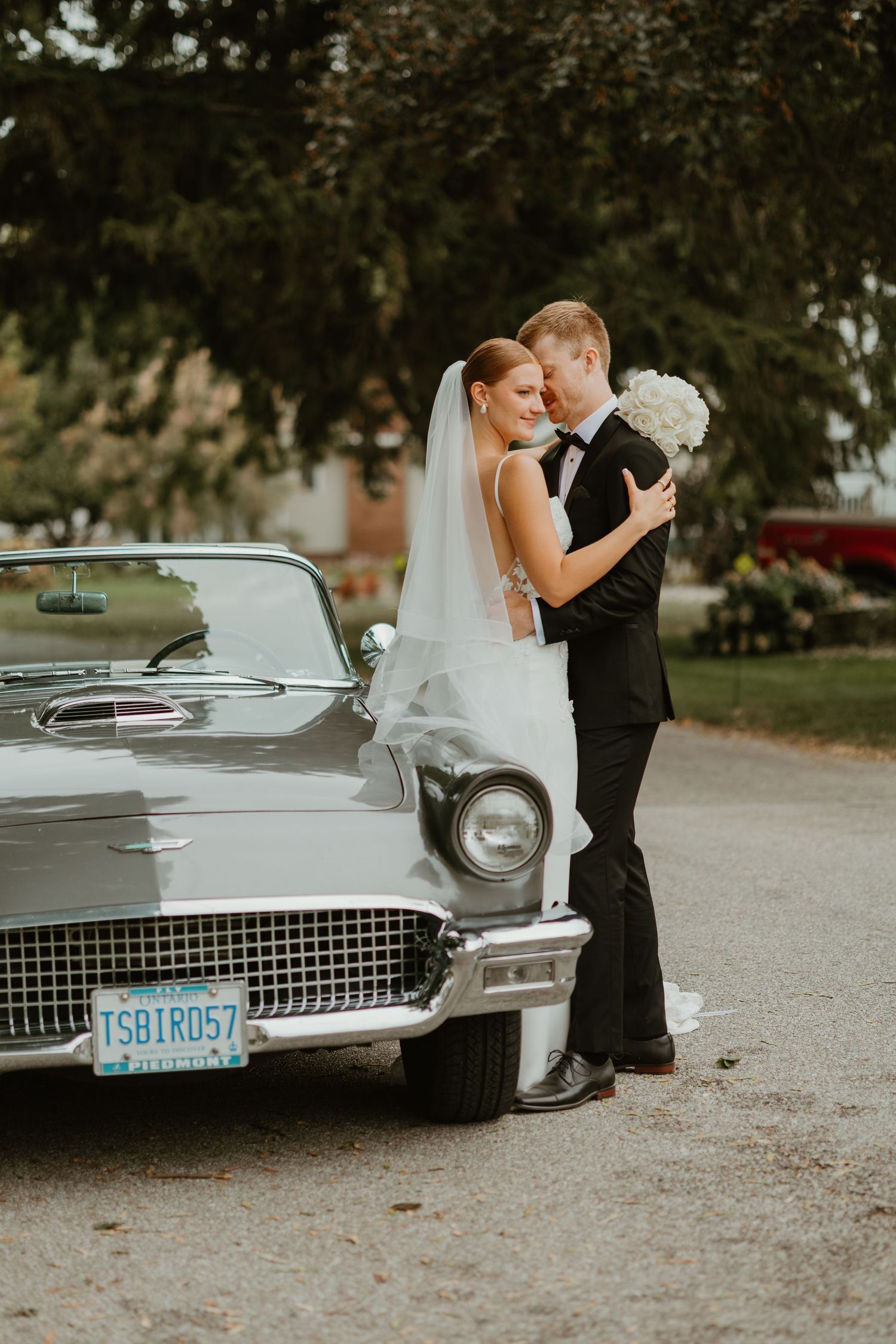 Bride and groom embracing beside a classic vintage Ford Thunderbird convertible on their wedding day.