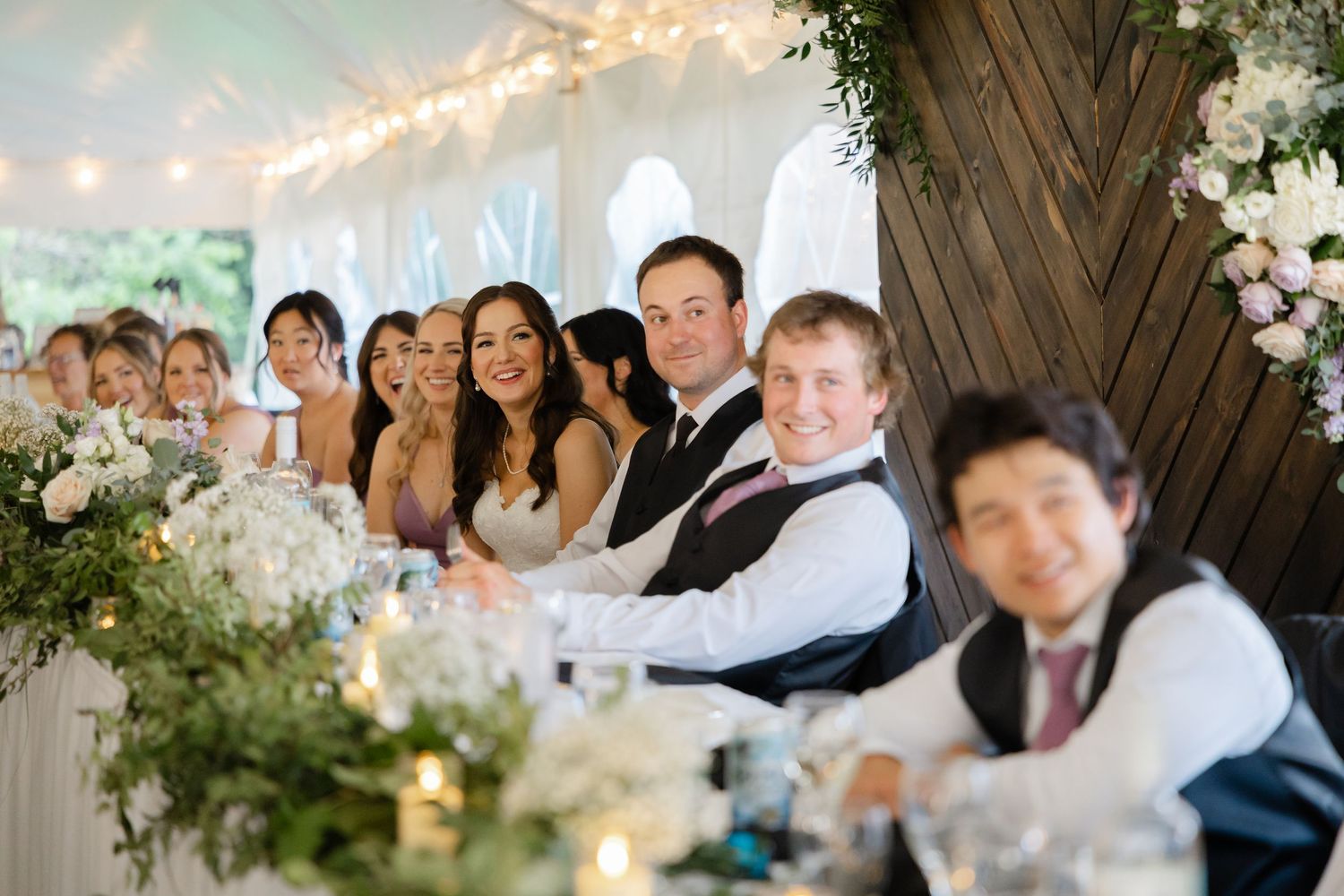Bride, groom, and wedding party smile and laugh at decorated head table during elegant tented wedding reception.