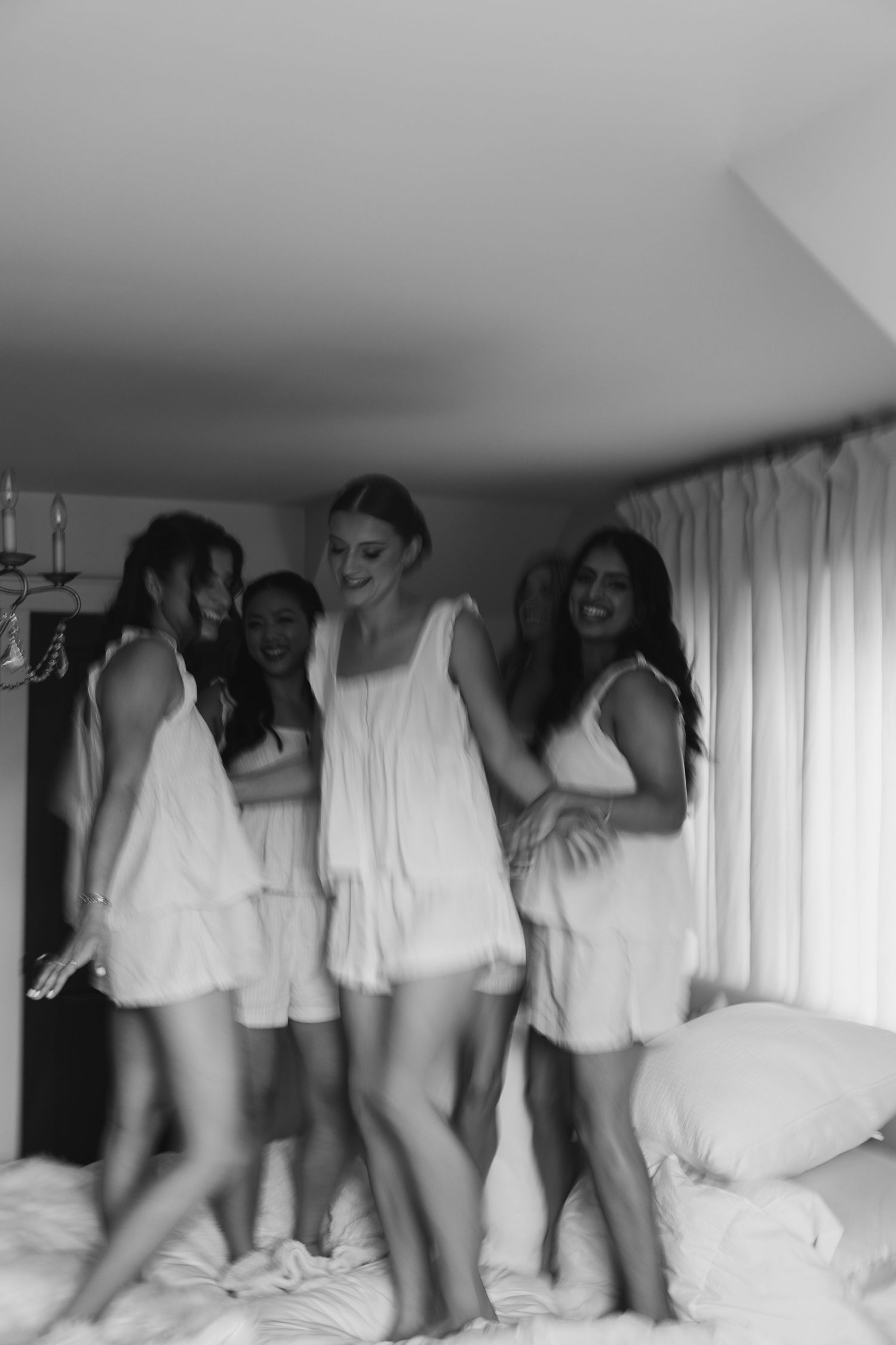 Black and white photo of bride and bridesmaids in white pajamas jumping on a bed together.
