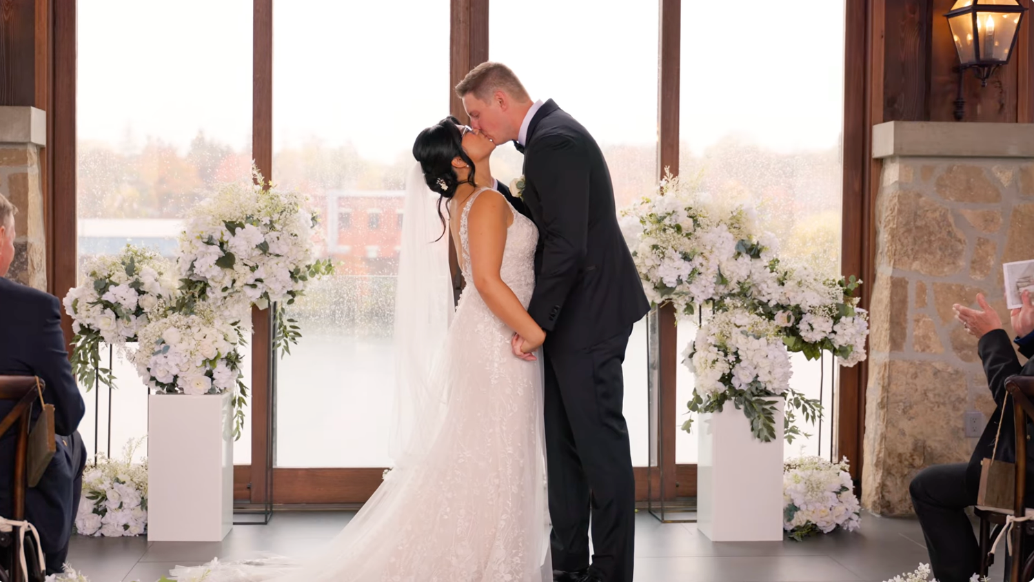 Bride and groom share first kiss at indoor wedding ceremony flanked by white floral arrangements near large windows.