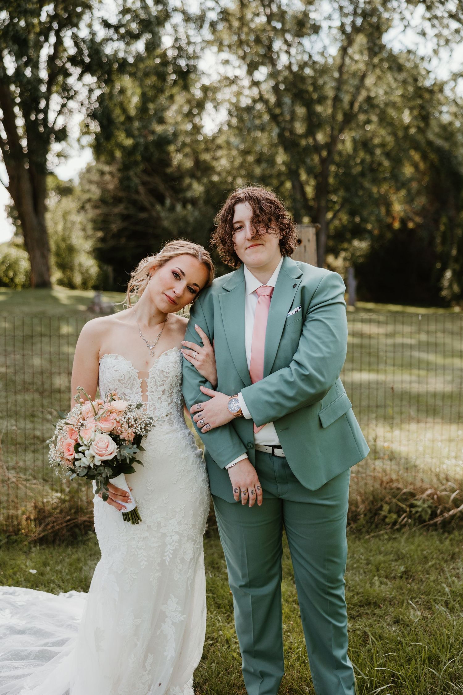 Bride in lace gown and partner in teal suit posing together in garden setting.