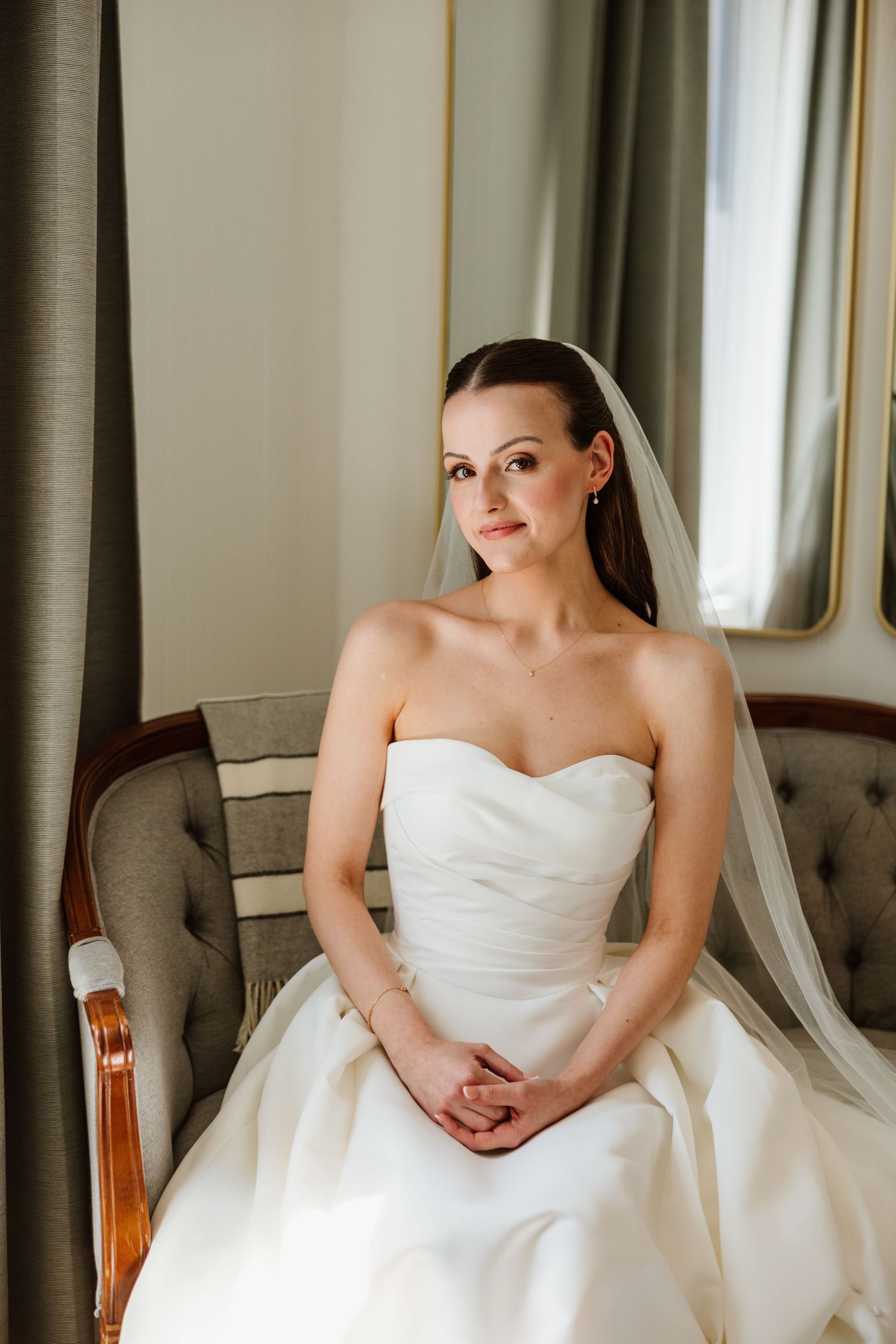 Elegant bride in strapless gown and veil sits by window in soft natural light.