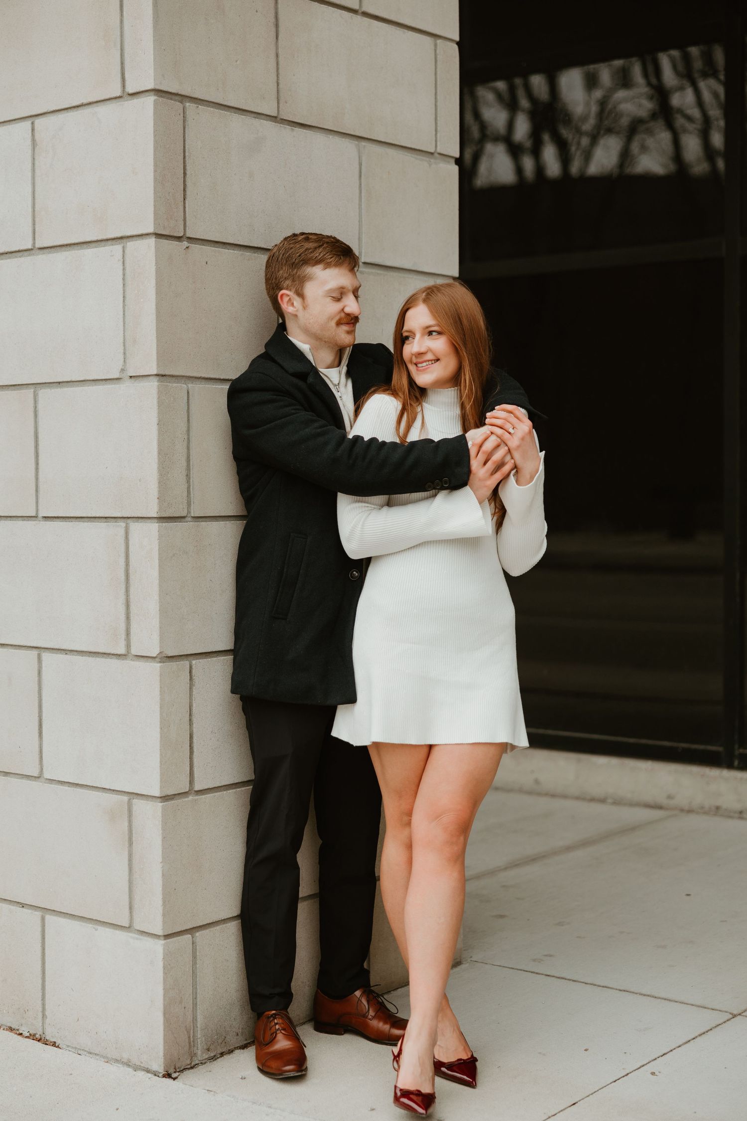 Man in black suit embraces smiling woman in white mini dress against a stone column outdoors.