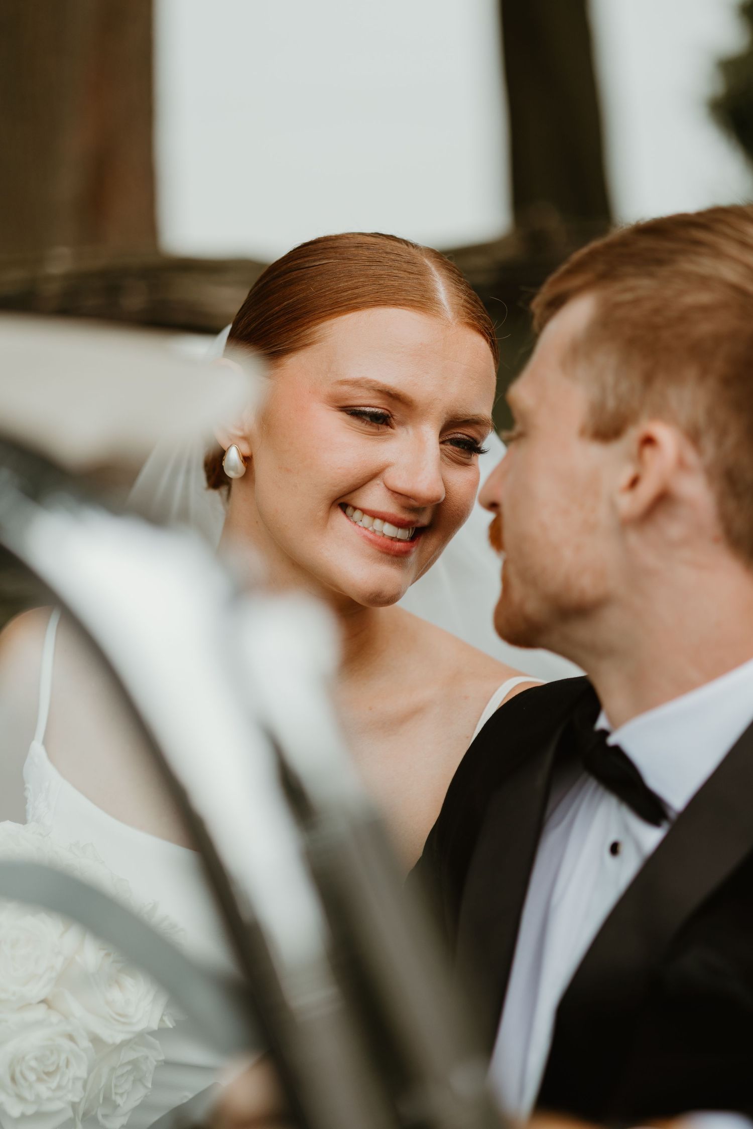 Bride smiling at groom in black tuxedo during romantic wedding portrait near vintage car.