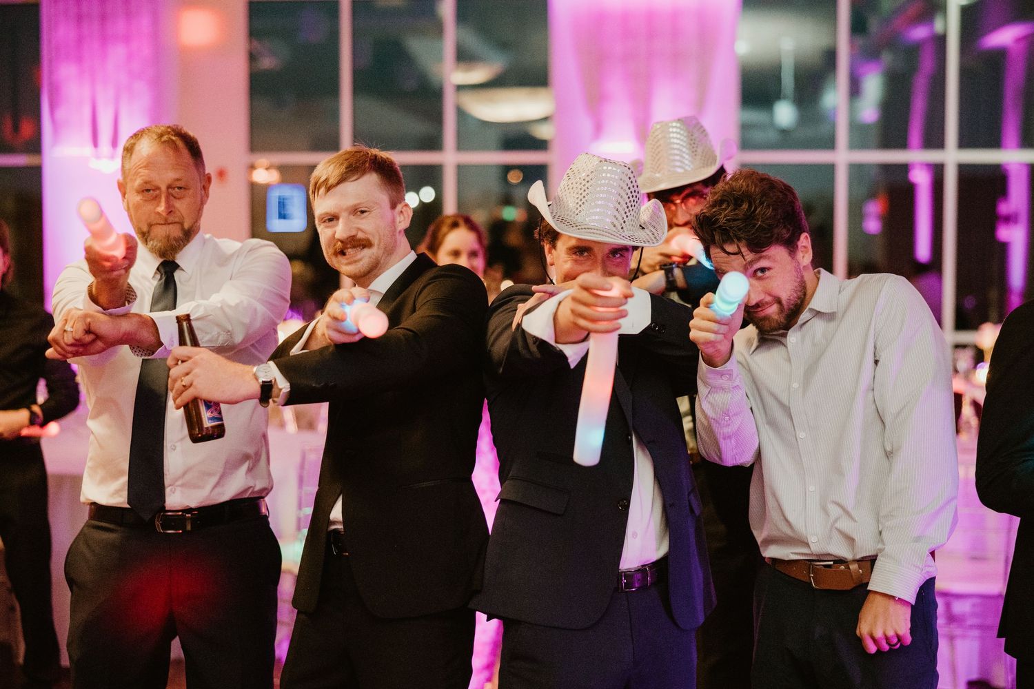 Group of men in suits strike a fun pose pointing finger guns at the camera during a wedding reception.