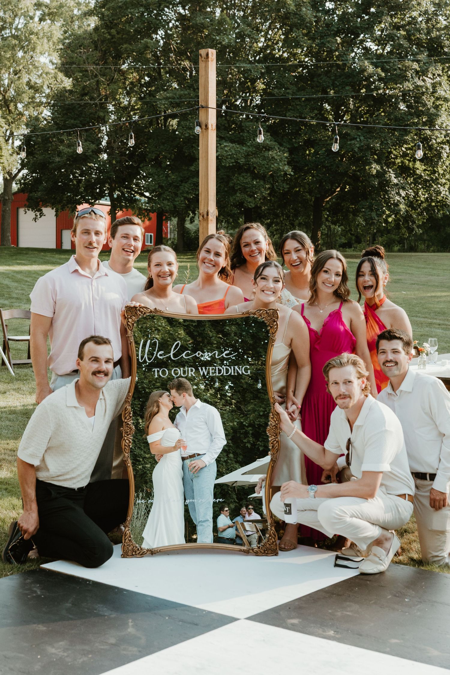 Wedding party posing with a decorative welcome sign at an outdoor summer wedding.