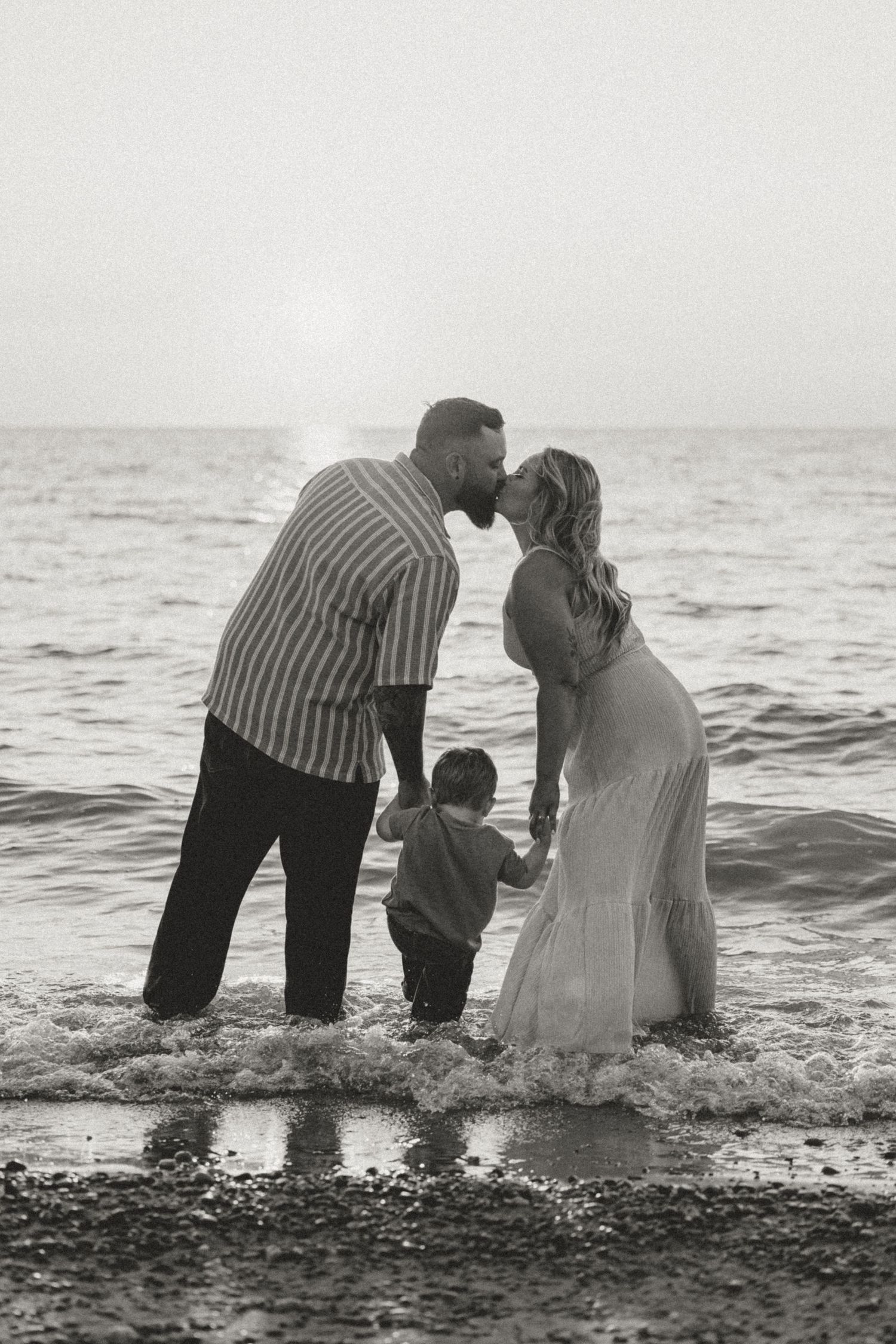 Black and white photo of couple kissing at beach shoreline while young child stands between them in ocean waves.