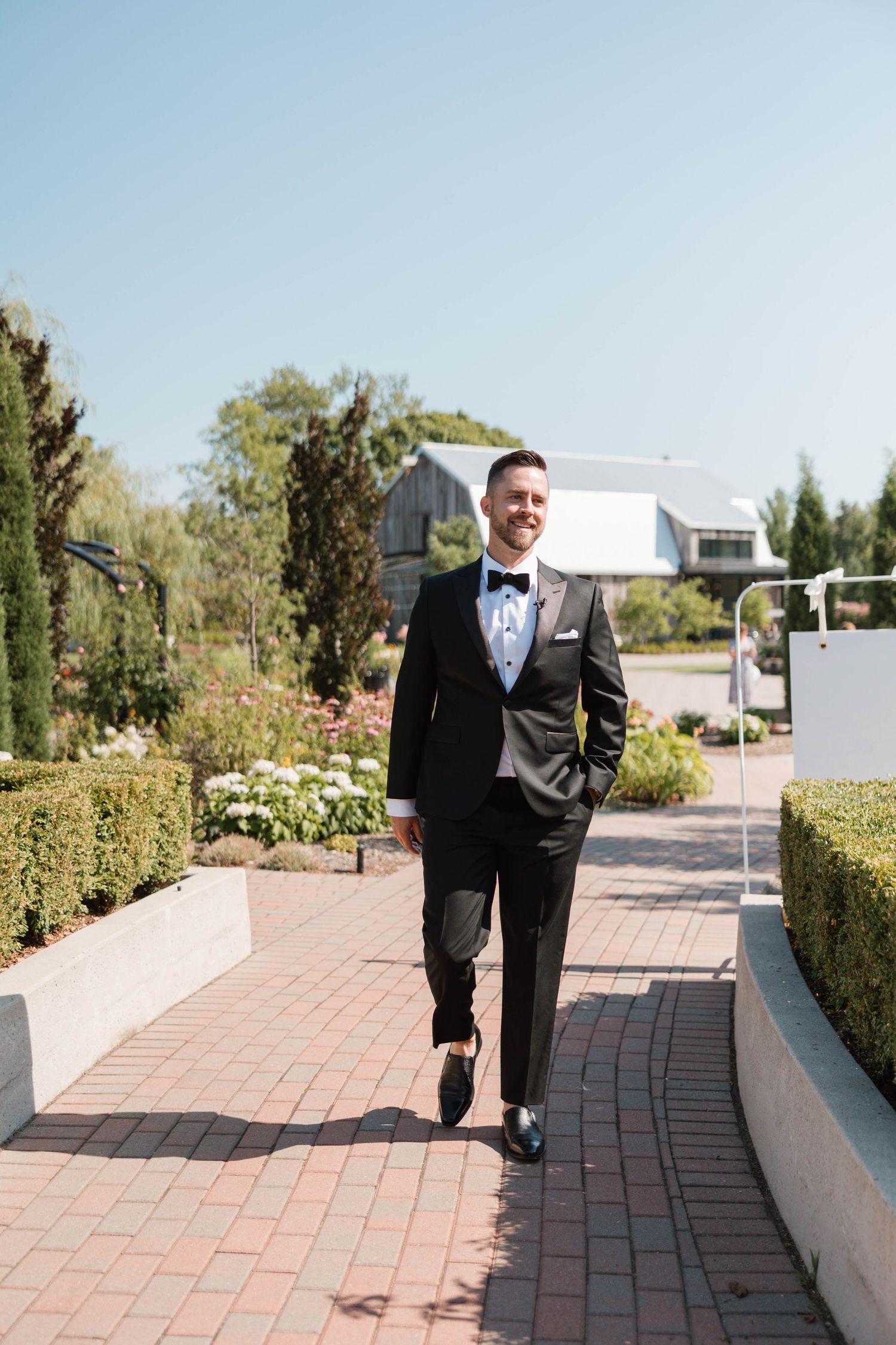 Groom in black tuxedo walking confidently along a sunny garden brick pathway.