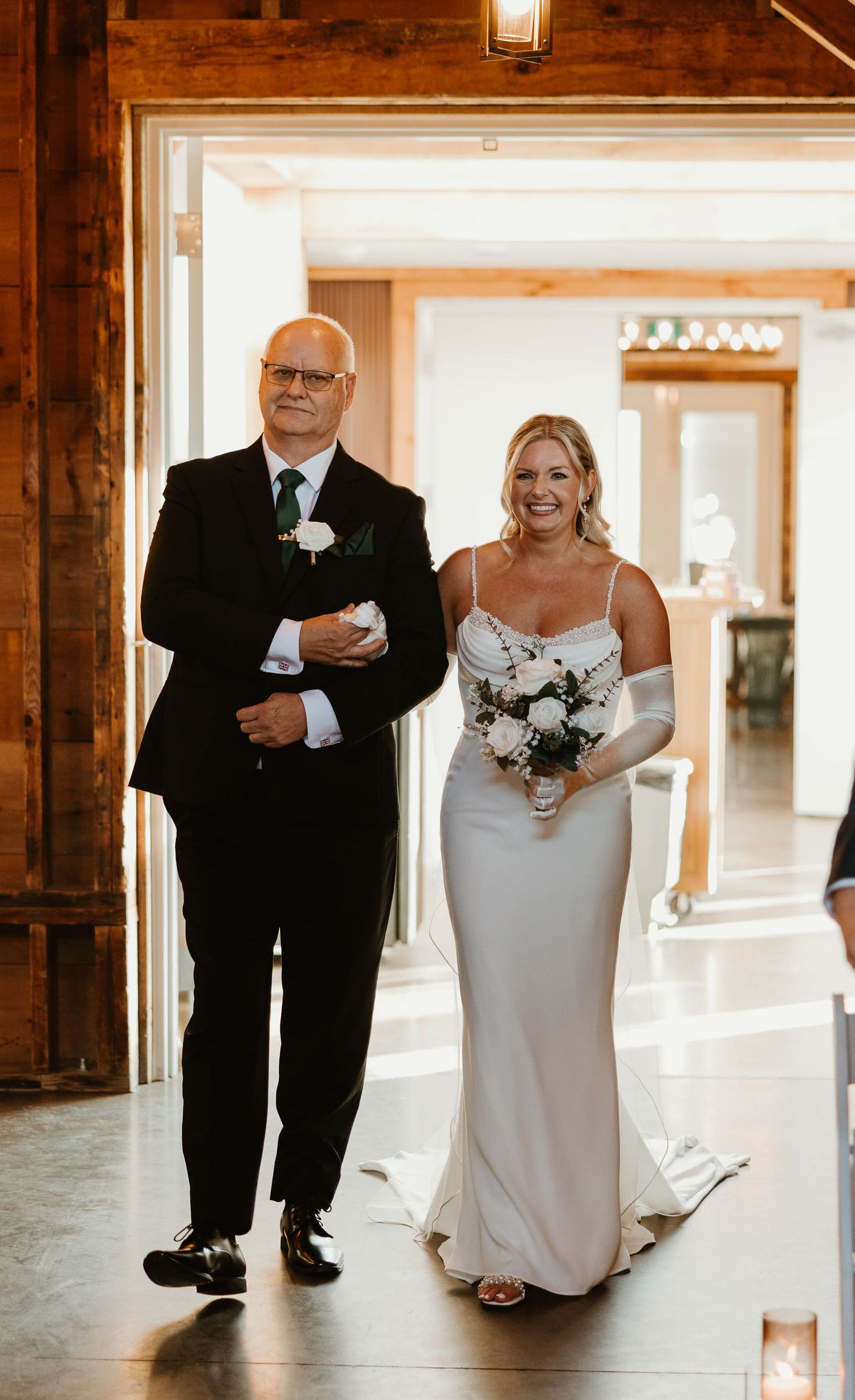 Smiling bride in white gown walks down aisle with father in black suit at rustic barn wedding.