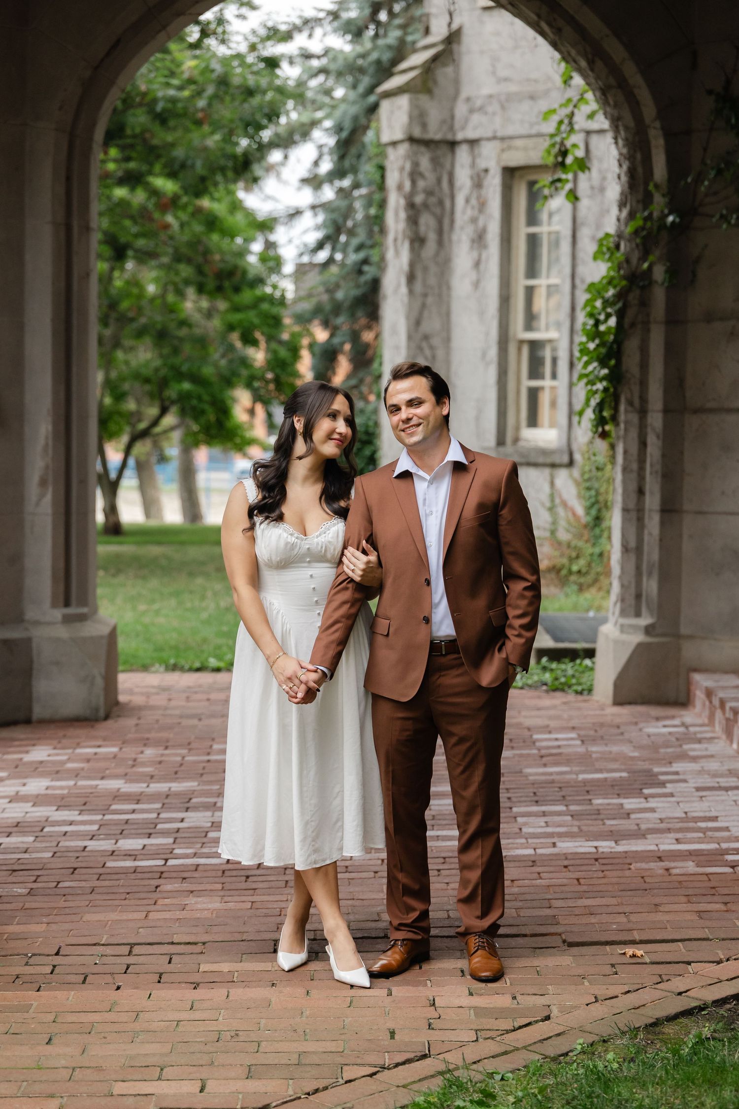 Couple holding hands and smiling under a stone archway on a brick path, she in a white dress and he in a brown suit, with trees behind.