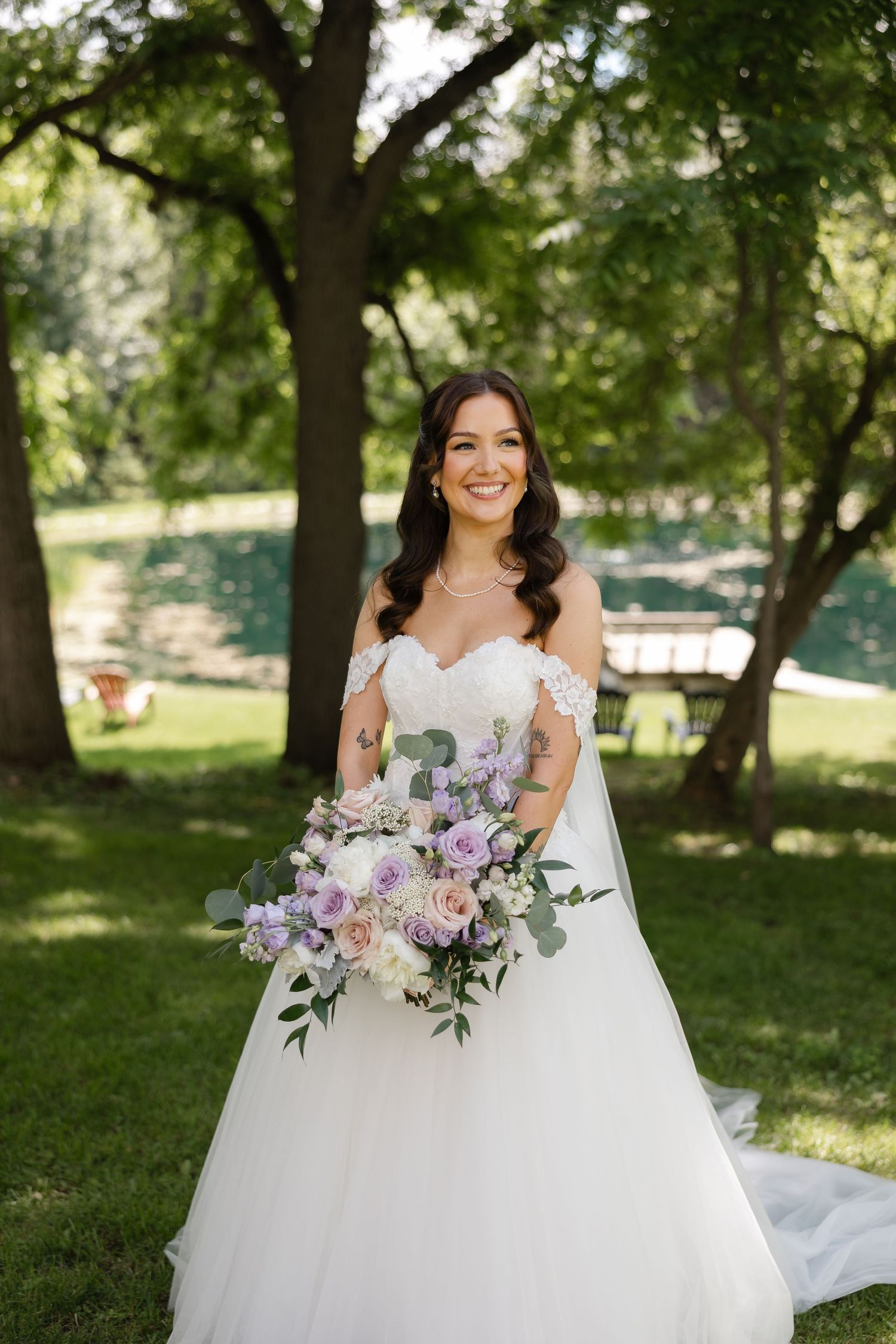 Radiant bride in off-shoulder white gown holds purple and white floral bouquet smiling outdoors near trees.