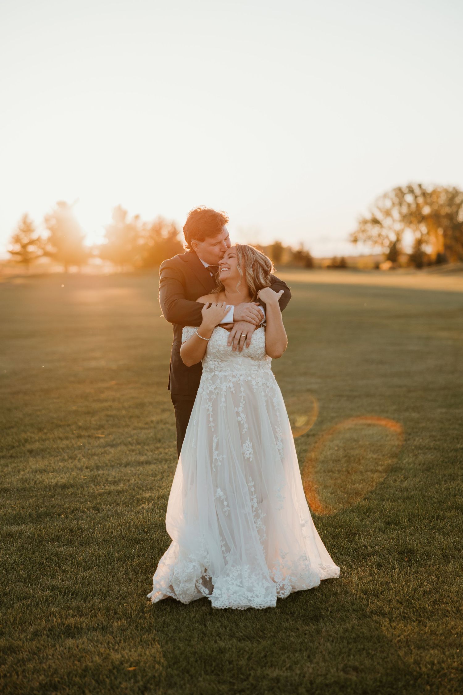 Bride in floral lace gown and groom embrace during a stunning golden hour sunset on a green field.