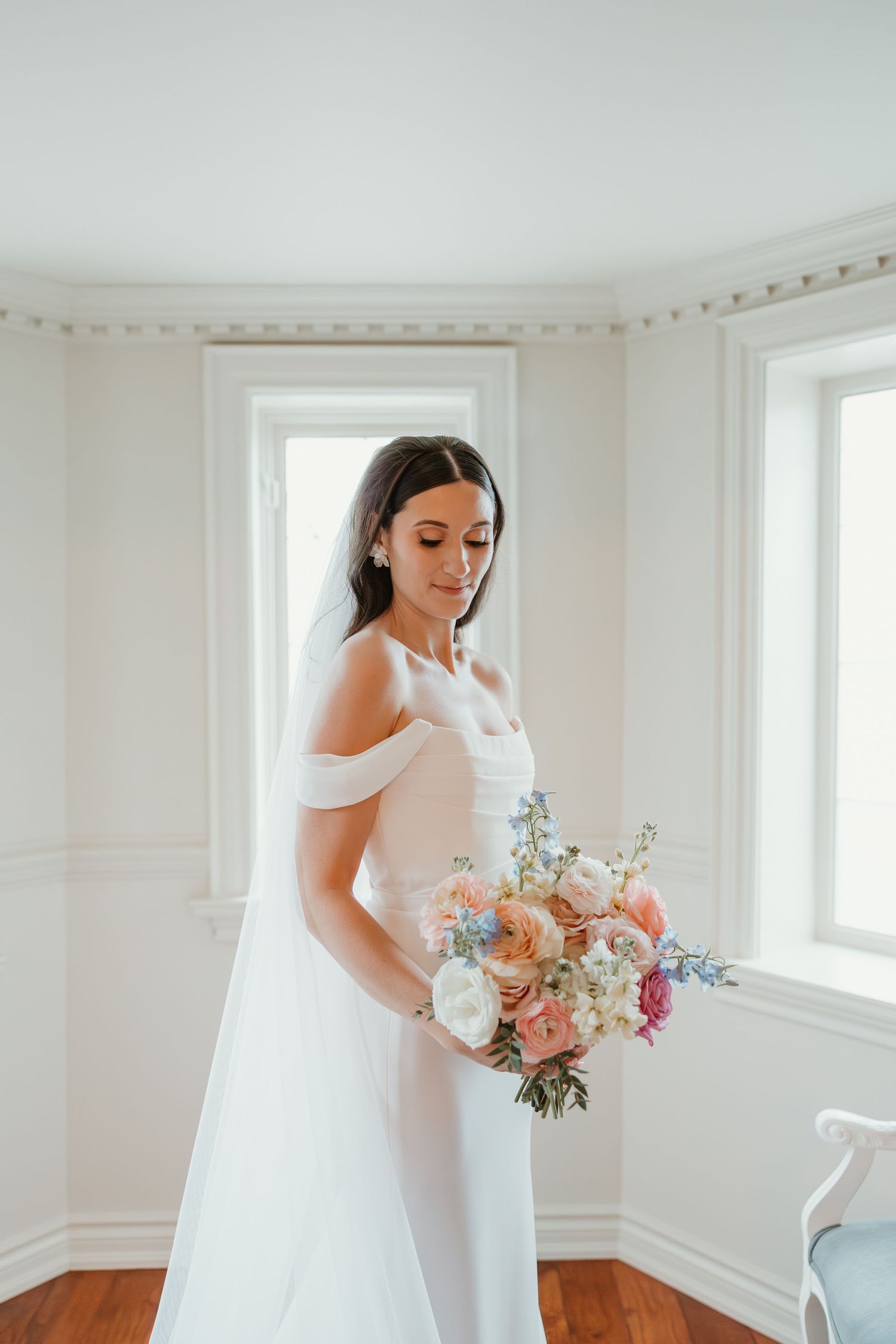 Bride in off-shoulder white gown holds a pastel bouquet of roses and wildflowers in a bright room.