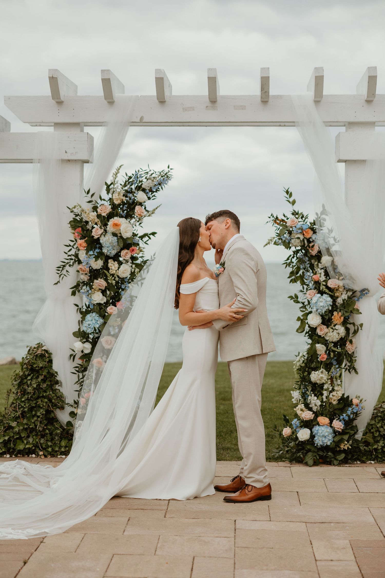 Bride and groom sharing first kiss under a white pergola adorned with pastel flowers overlooking the ocean.