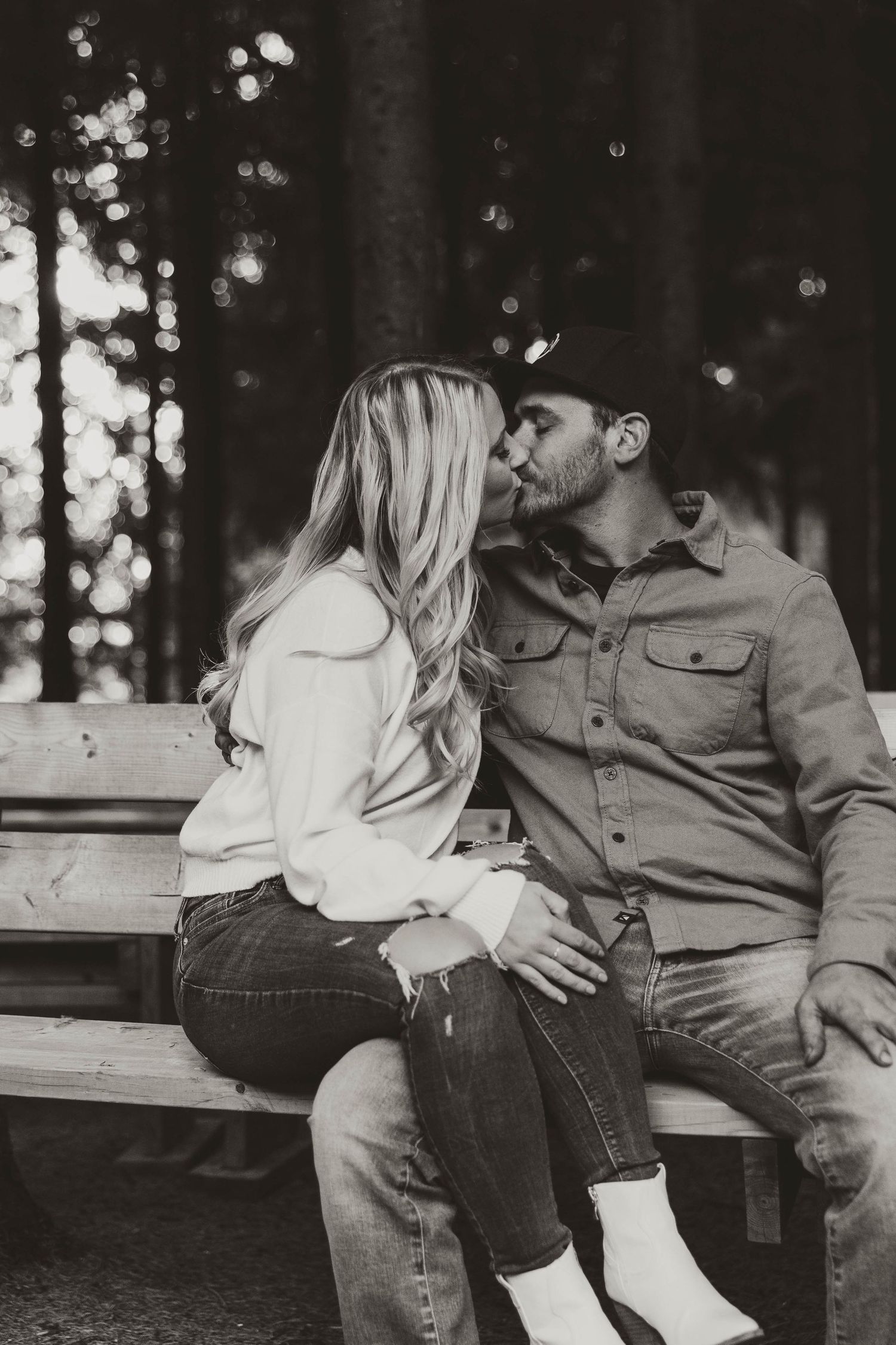 Black and white photo of couple kissing on wooden bench in forest setting.